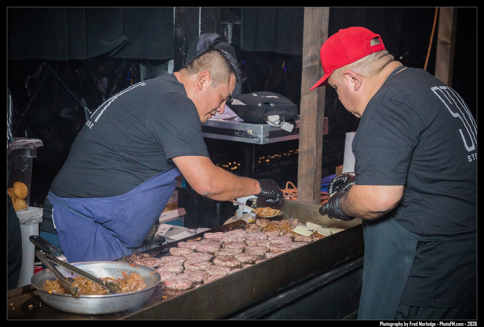 Two men preparing meat at a food stall or outdoor kitchen, with one adding sauce or marinade to patties and the other assisting, both wearing black shirts and black gloves.