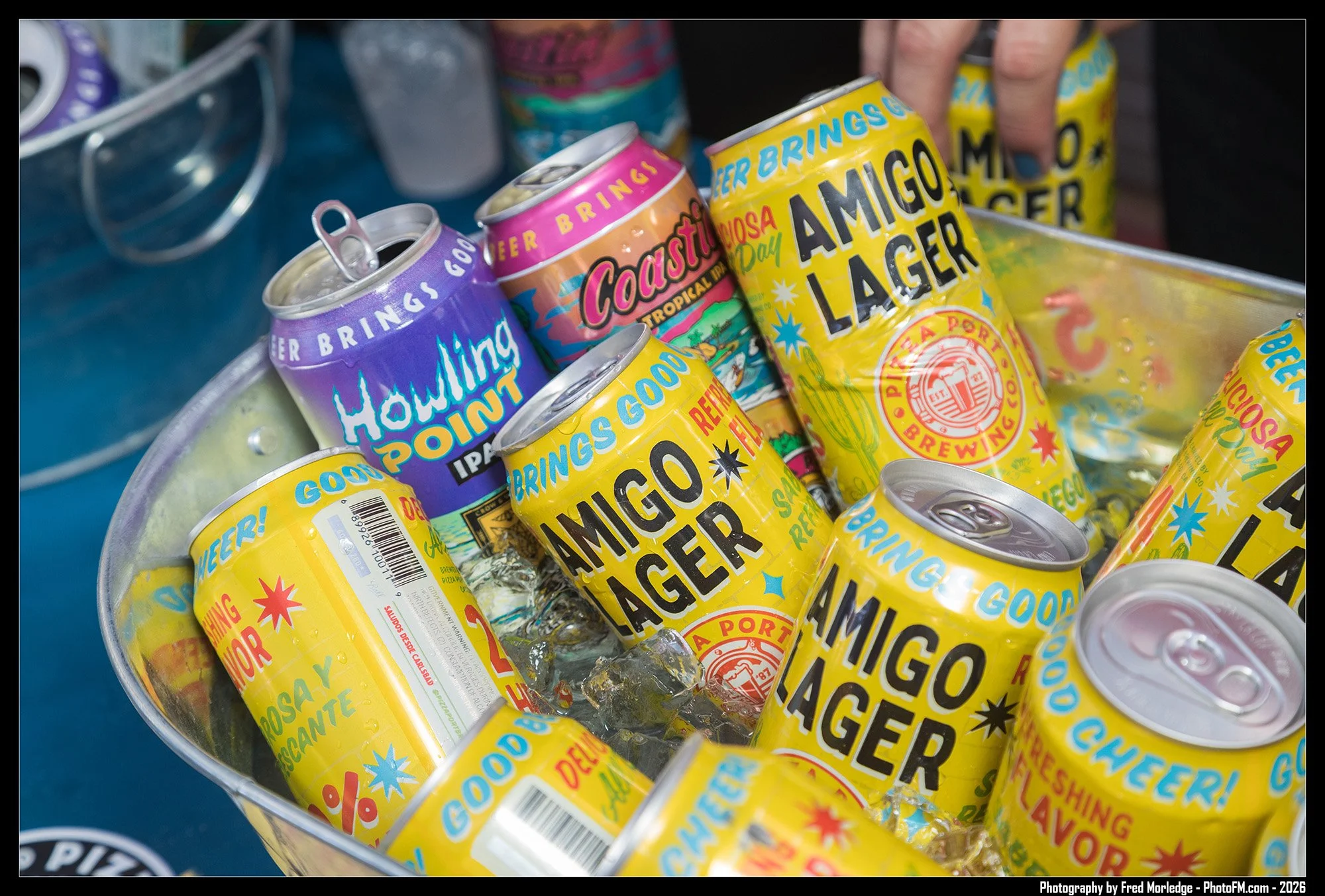 A metal bowl filled with ice and opened cans of beer with colorful labels, including yellow, purple, and pink, on a blue surface.