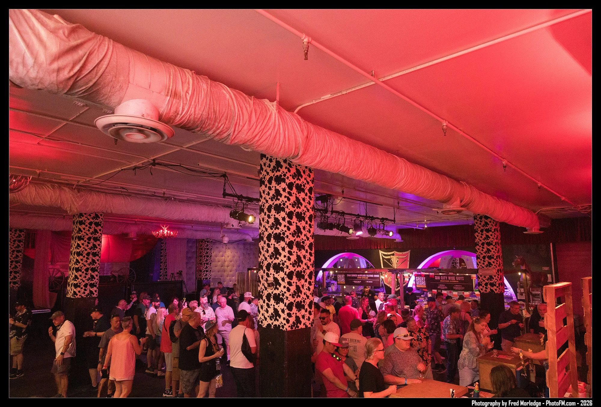 Crowd of people at an indoor event or festival with decorated speakers and a black and white spotted column, illuminated by pink and purple lighting.