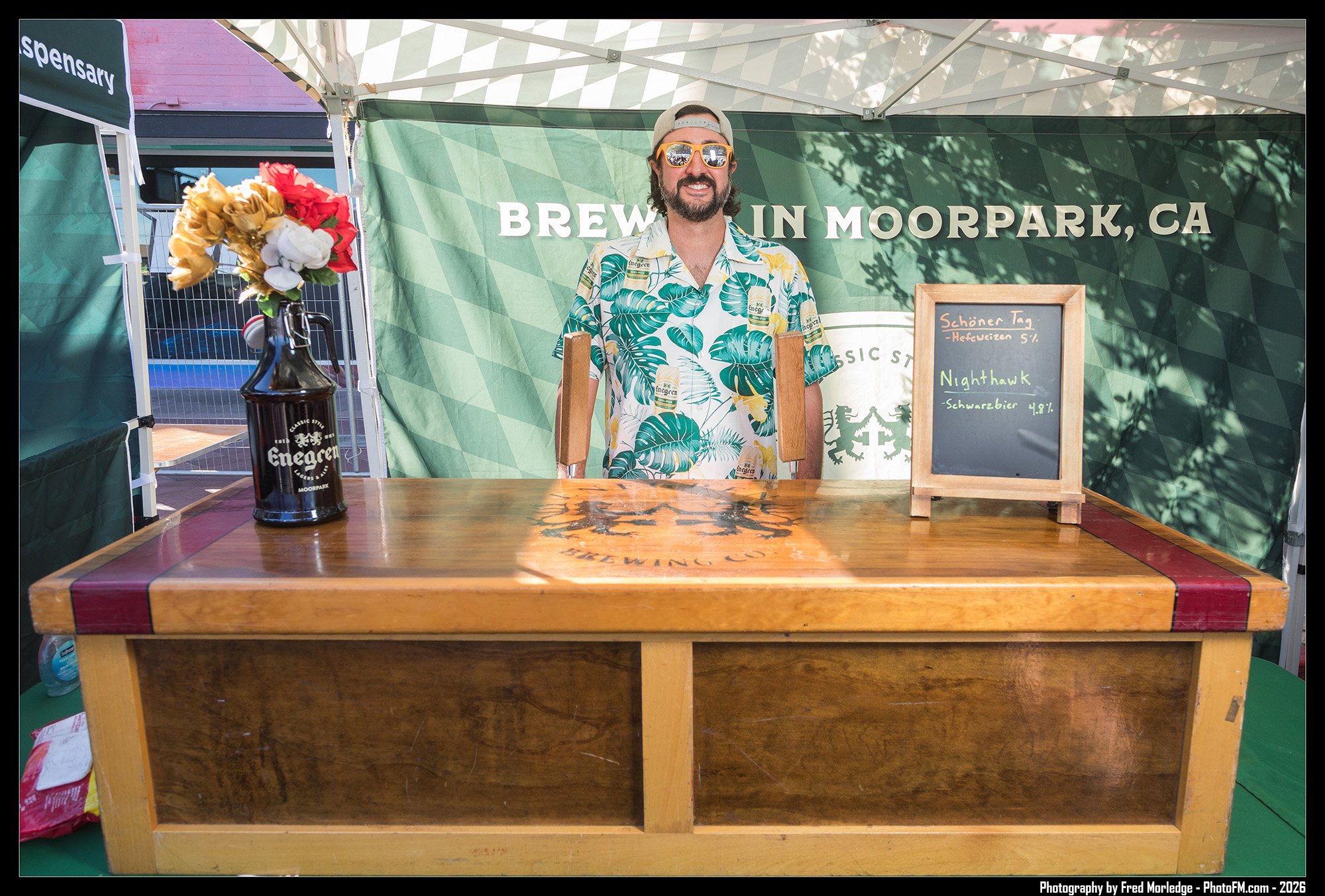 At an outdoor booth with a green backdrop that says 'Brew in Moorpark, CA', a man wearing a floral Hawaiian shirt, sunglasses, and a cap is smiling. A small chalkboard menu lists beers like 'Nighthawk' and 'Schöner Tag'. A table in front has a bottle