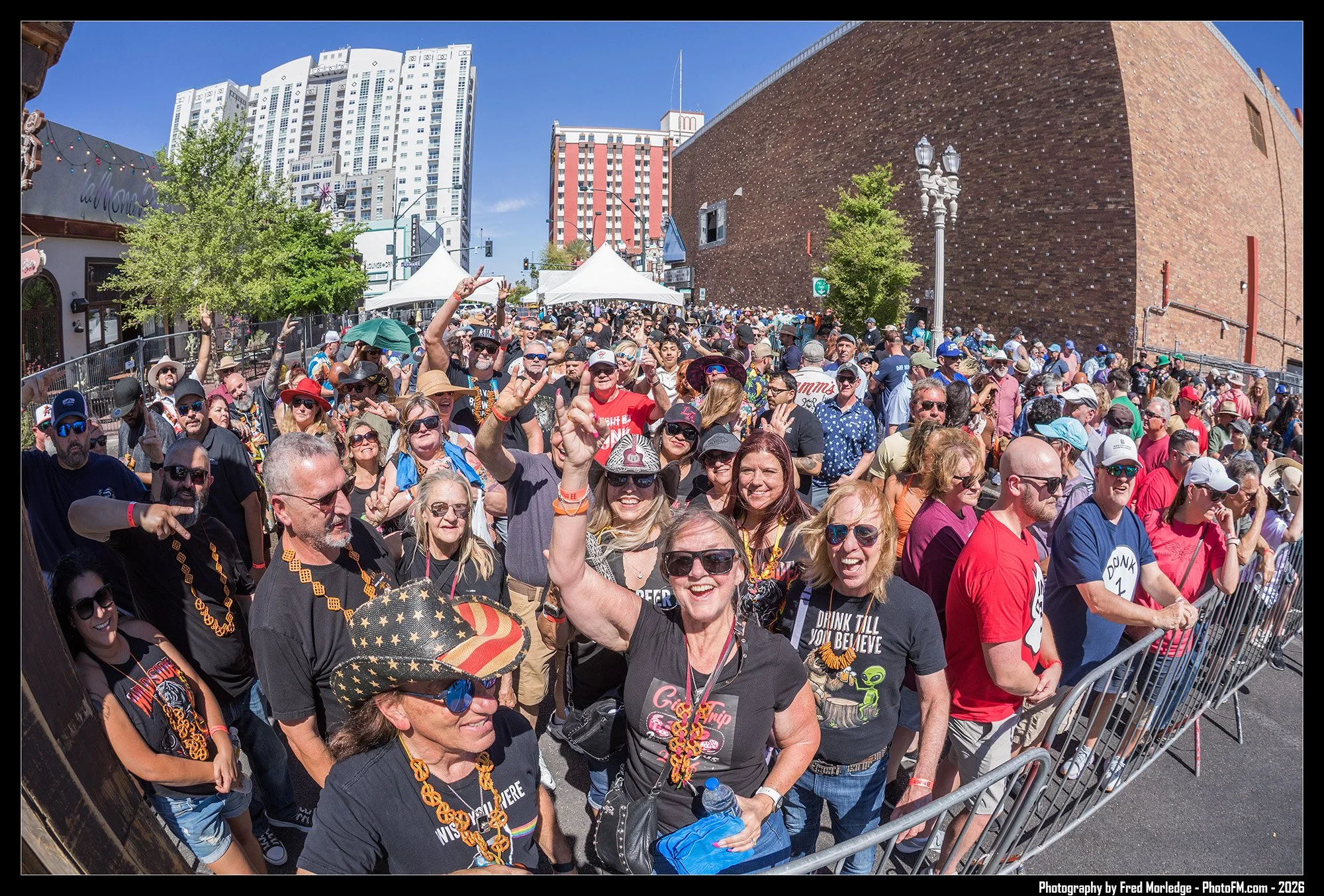 Crowd of people gathered outdoors at an event on a sunny day, with tents, trees, and tall buildings in the background.