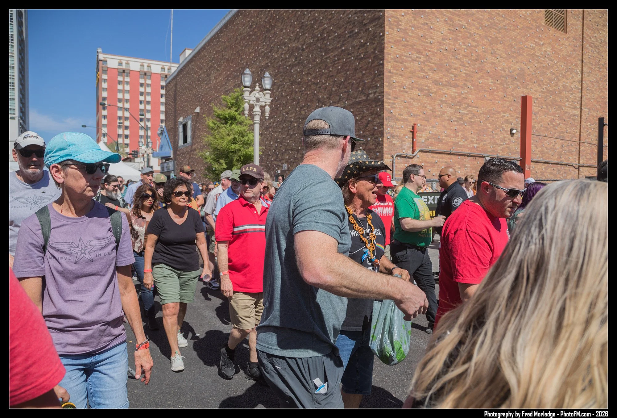 Crowd of people walking outdoors on a sunny day in an urban area with brick buildings, trees, and street lamps in the background.