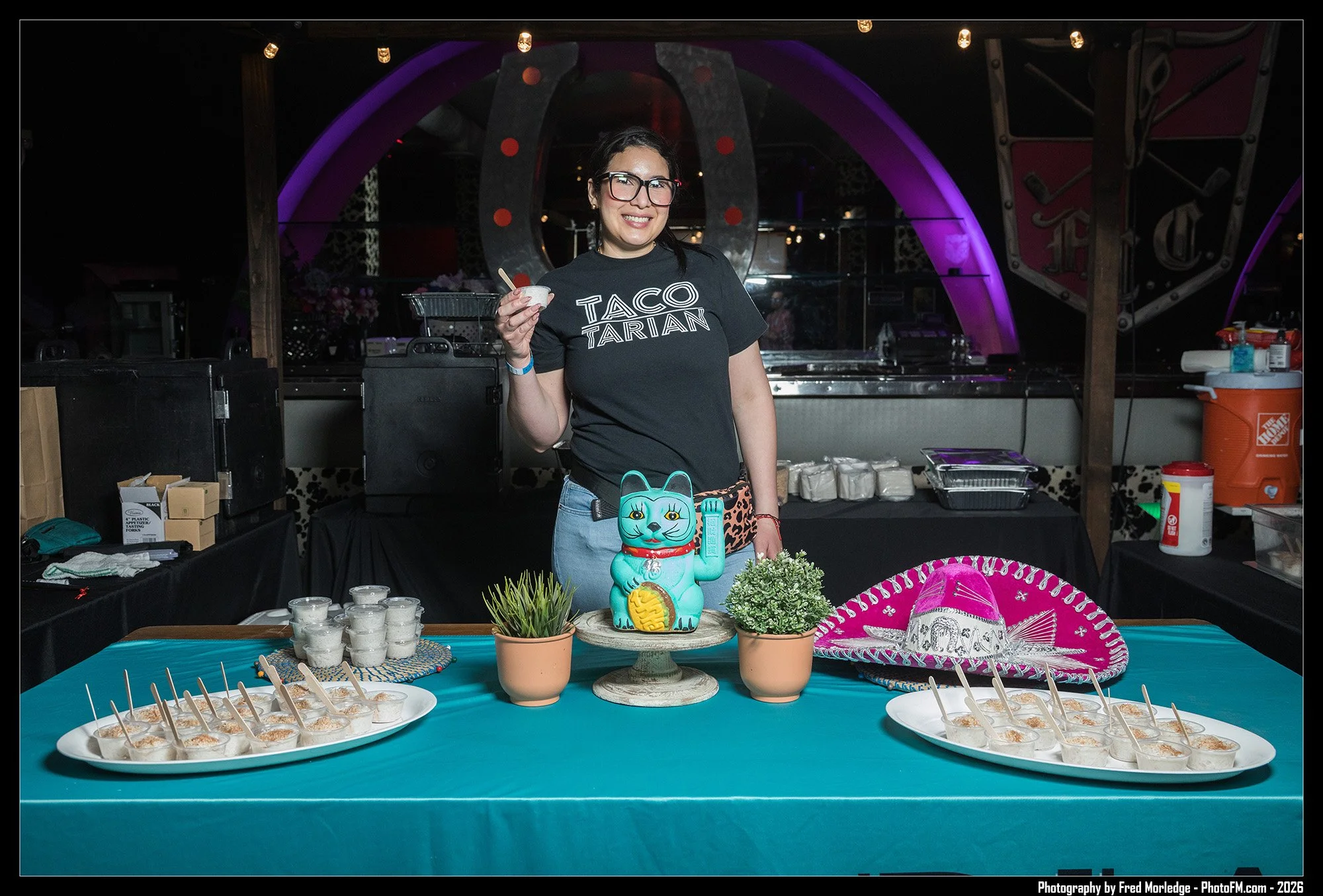 A woman wearing glasses and a black T-shirt with the words 'Taco Tarian' standing behind a table with Mexican-themed decorations and food. She is holding a small bowl and smiling. The table has several small cups of a dessert with sticks and a decora