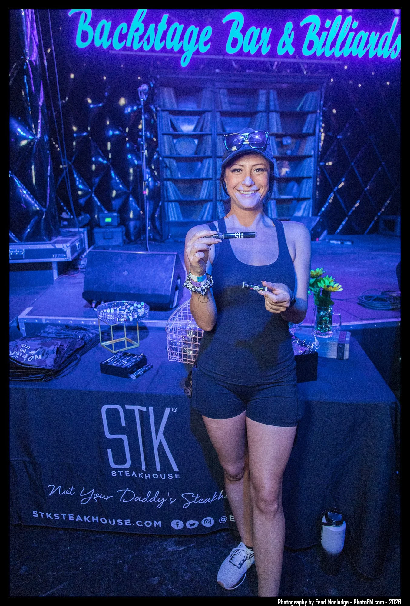A woman smiling and holding a marker pen inside a music event venue, with a backdrop that reads 'Backstage Bar & Billiards' and 'STK Steakhouse'. She is wearing a black tank top, black shorts, white sneakers, a cap, glasses on her head, and a wristba