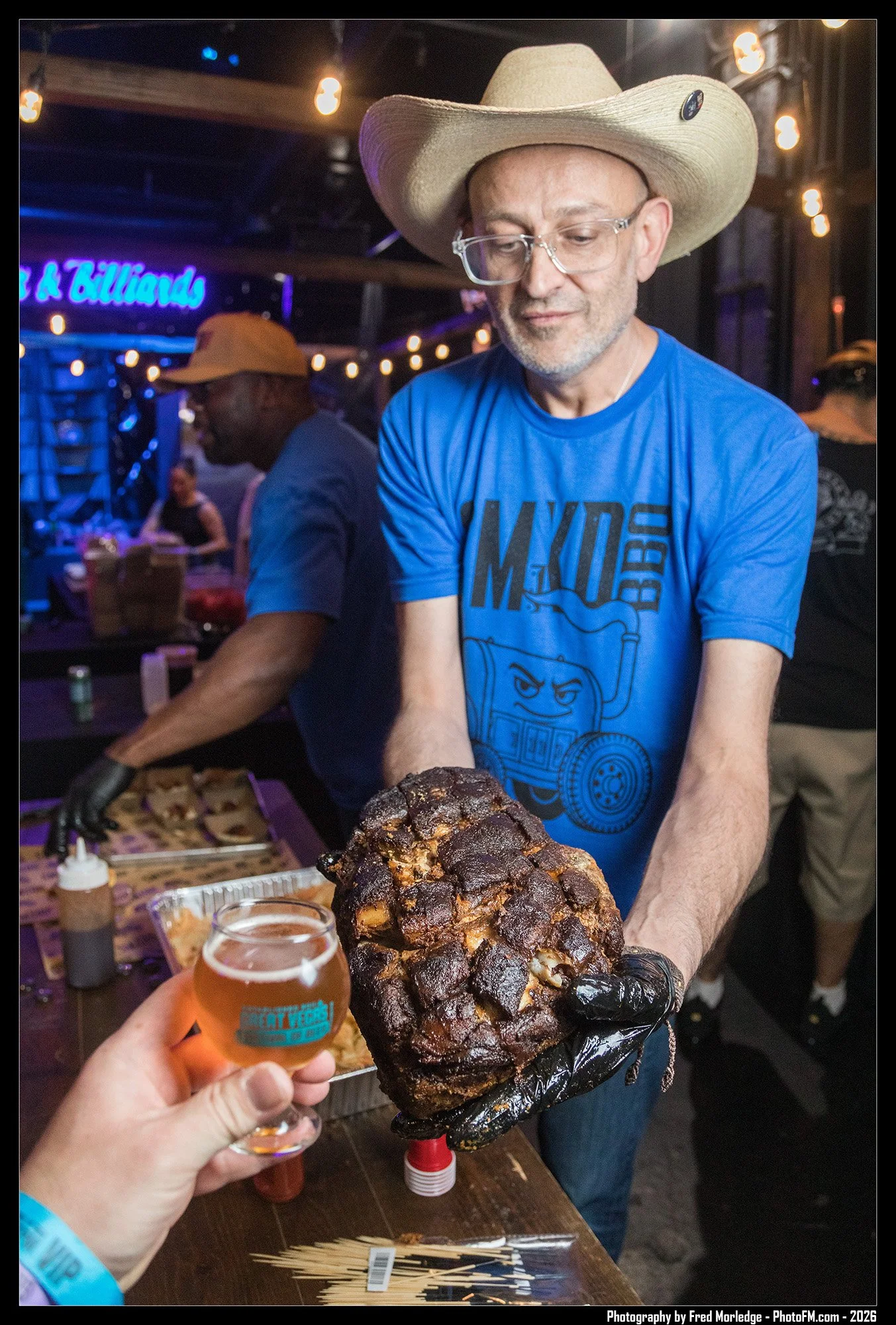 A man wearing a cowboy hat and glasses is serving grilled meat at a social gathering. In the foreground, a hand is holding a glass of beer, and in the background, there are other people and a sign that reads '~illions' in neon lights.