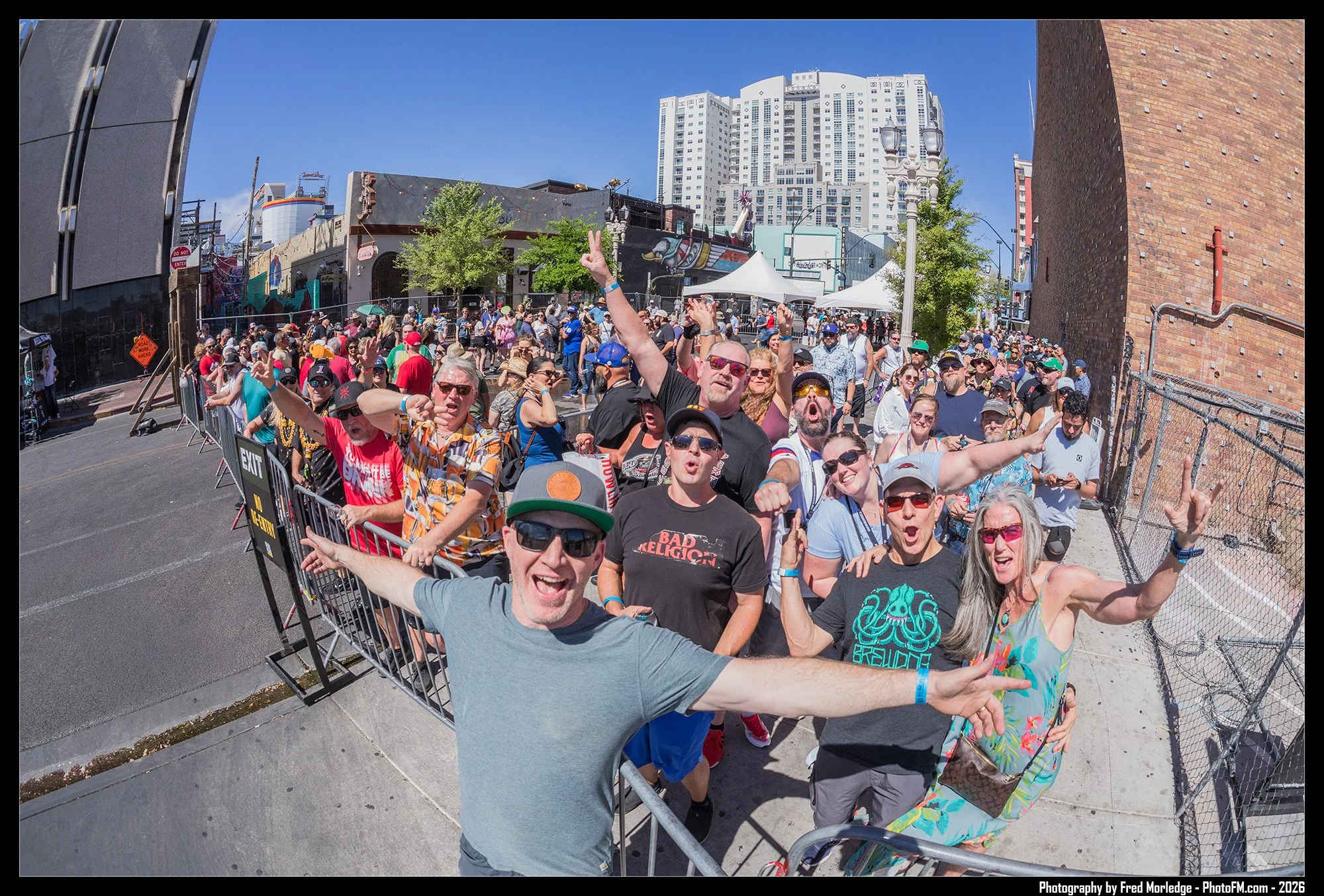 Crowd of people standing behind a metal barricade at an outdoor event, with some smiling and making peace signs, under a clear blue sky.