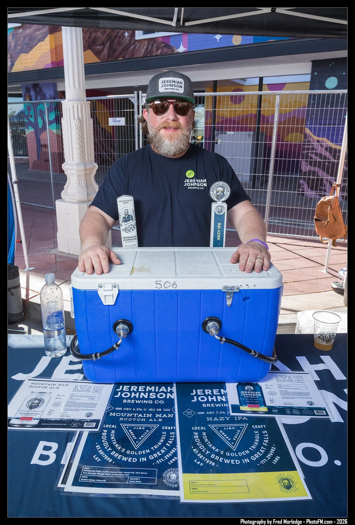 A man with sunglasses, a beard, and a dark t-shirt is standing behind a table displaying beer labels and information for Jeremiah Johnson Brewing Co., with a colorful mural in the background. He is holding a blue cooler with taps connected to it. On 