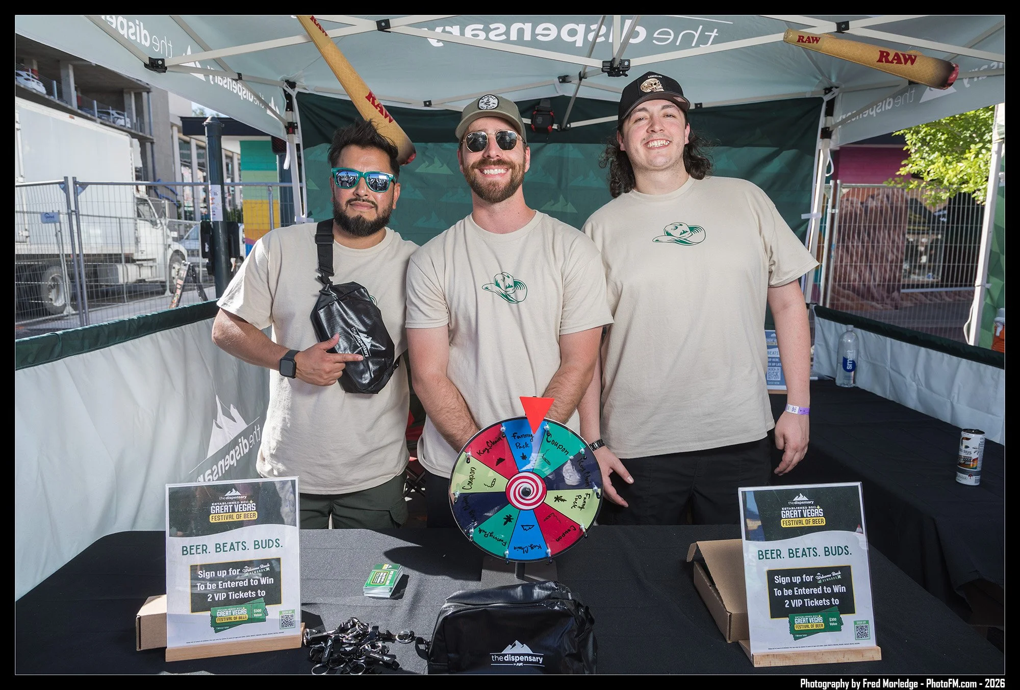 Three men stand under a tent at a festival, smiling at the camera. They wear casual T-shirts, and one has sunglasses and a camera around his neck. A colorful wheel on a table in front of them has sections labeled with prizes, and there are signs prom