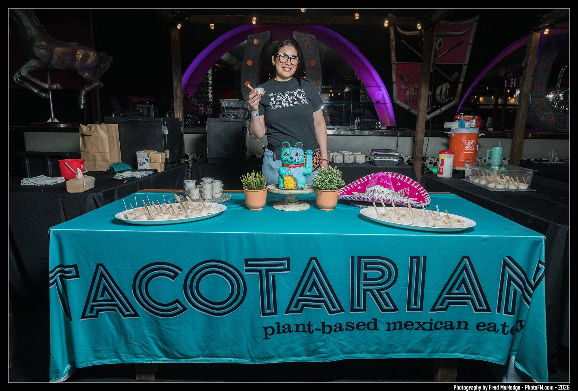 A woman standing behind a table with a blue tablecloth promoting plant-based Mexican eatery, Taco Tarian. She is holding a small cup, smiling, wearing a black T-shirt with 'TACO TARAN' printed on it, and glasses. The table has small cups with spoons 