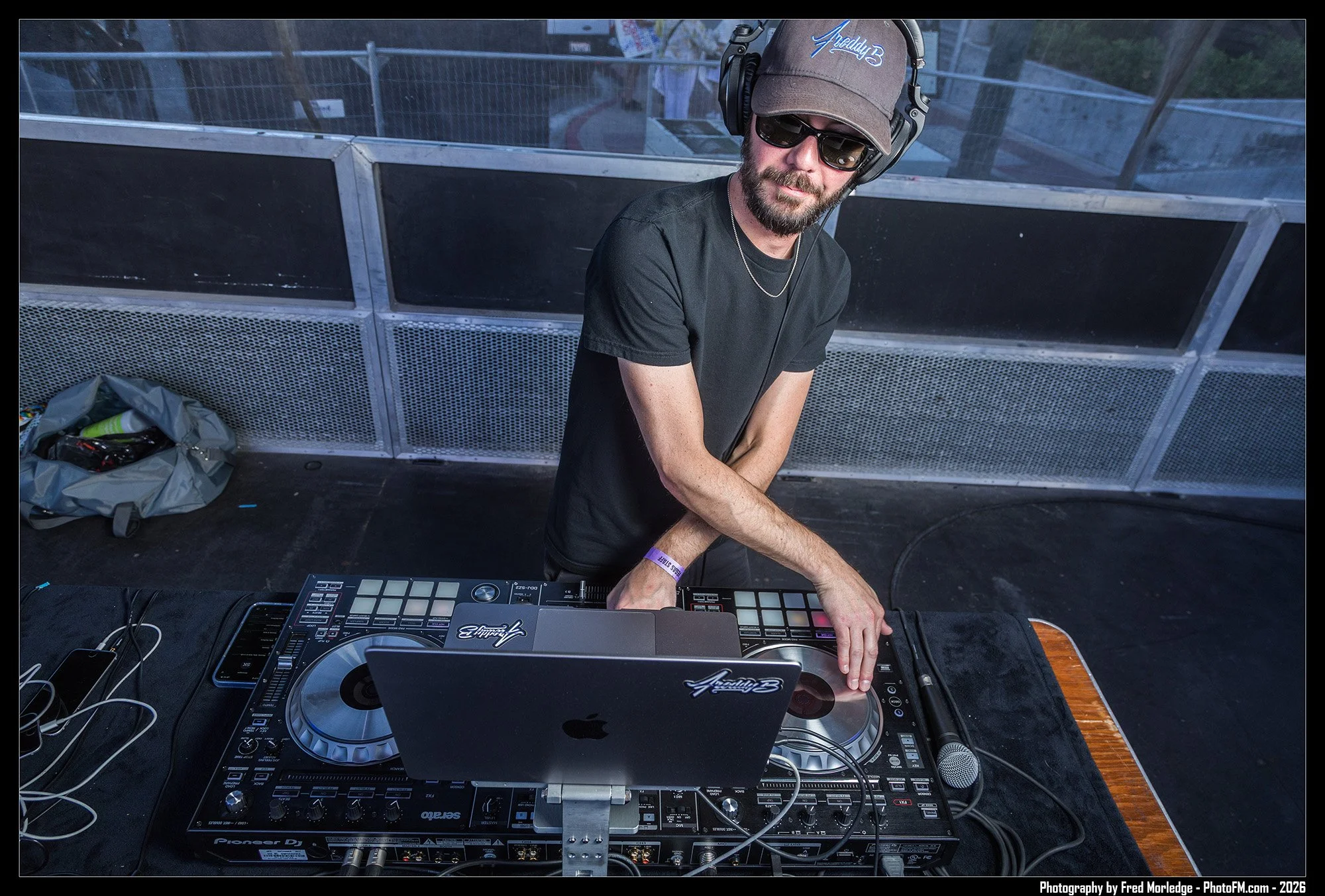 DJ wearing sunglasses, a cap, headphones, and a black shirt, DJing on a Pioneer DJ controller on a table, with a laptop, in an outdoor setting during daytime.