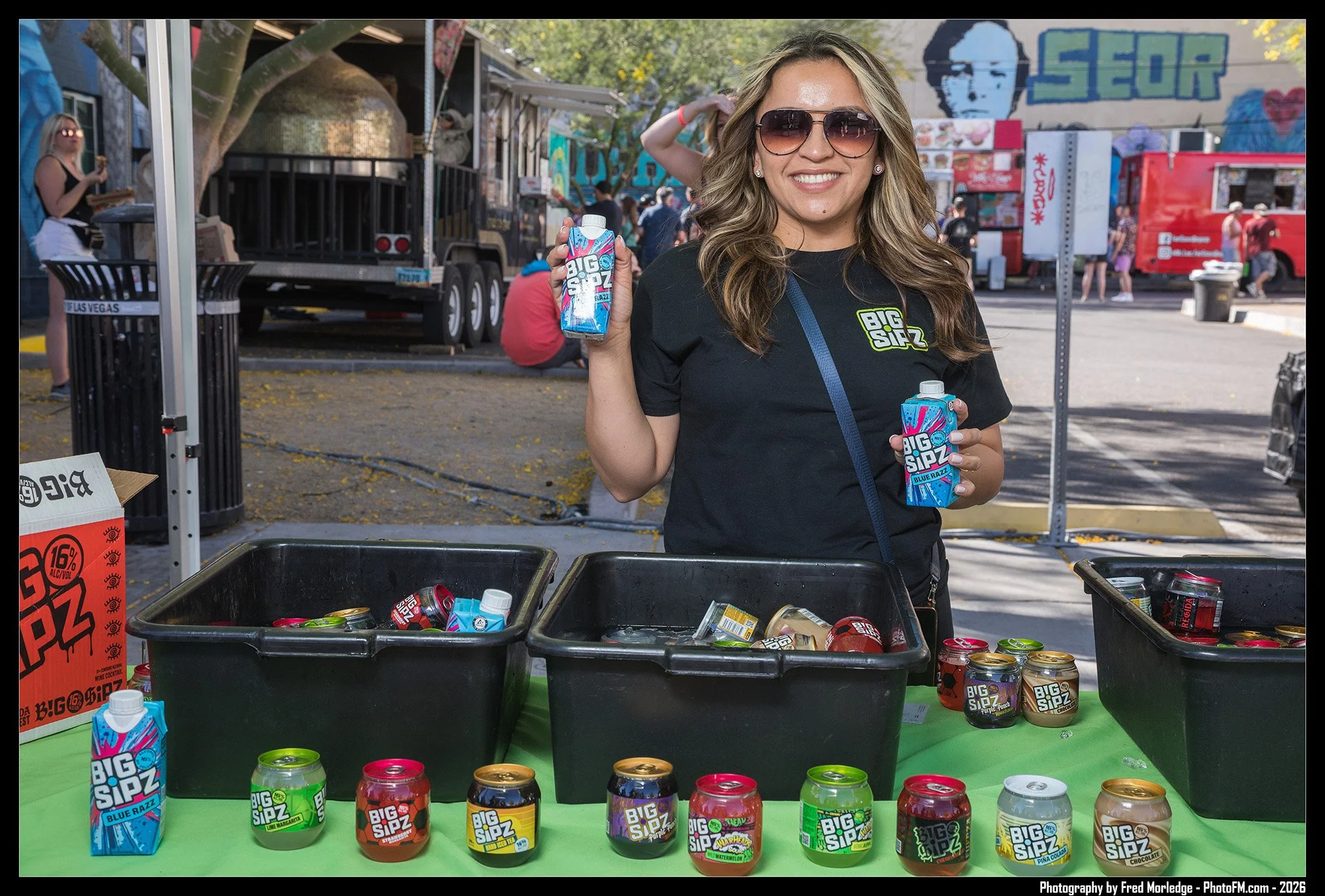 A woman wearing sunglasses and a black shirt with a 'BIG SIPZ' logo holds a juice box at an outdoor event, standing behind a table with various juice cans and boxes, with a food truck, people, and street art in the background.
