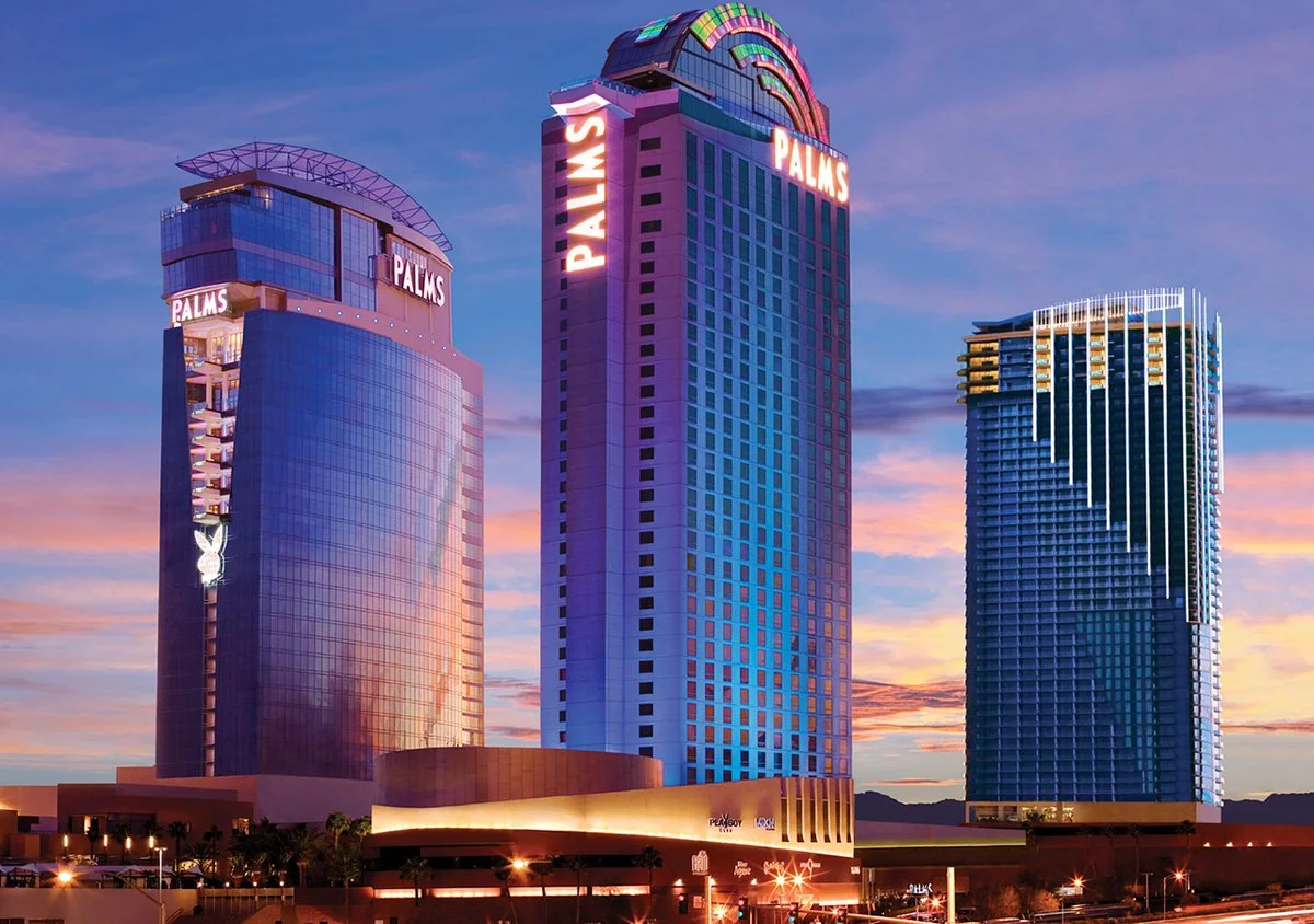 Nighttime view of three high-rise buildings in Las Vegas. The buildings are topped with colorful neon signs that say 'PALMS'. One building features the Playboy bunny logo. The sky has a mix of purple, pink, and orange hues from the sunset.