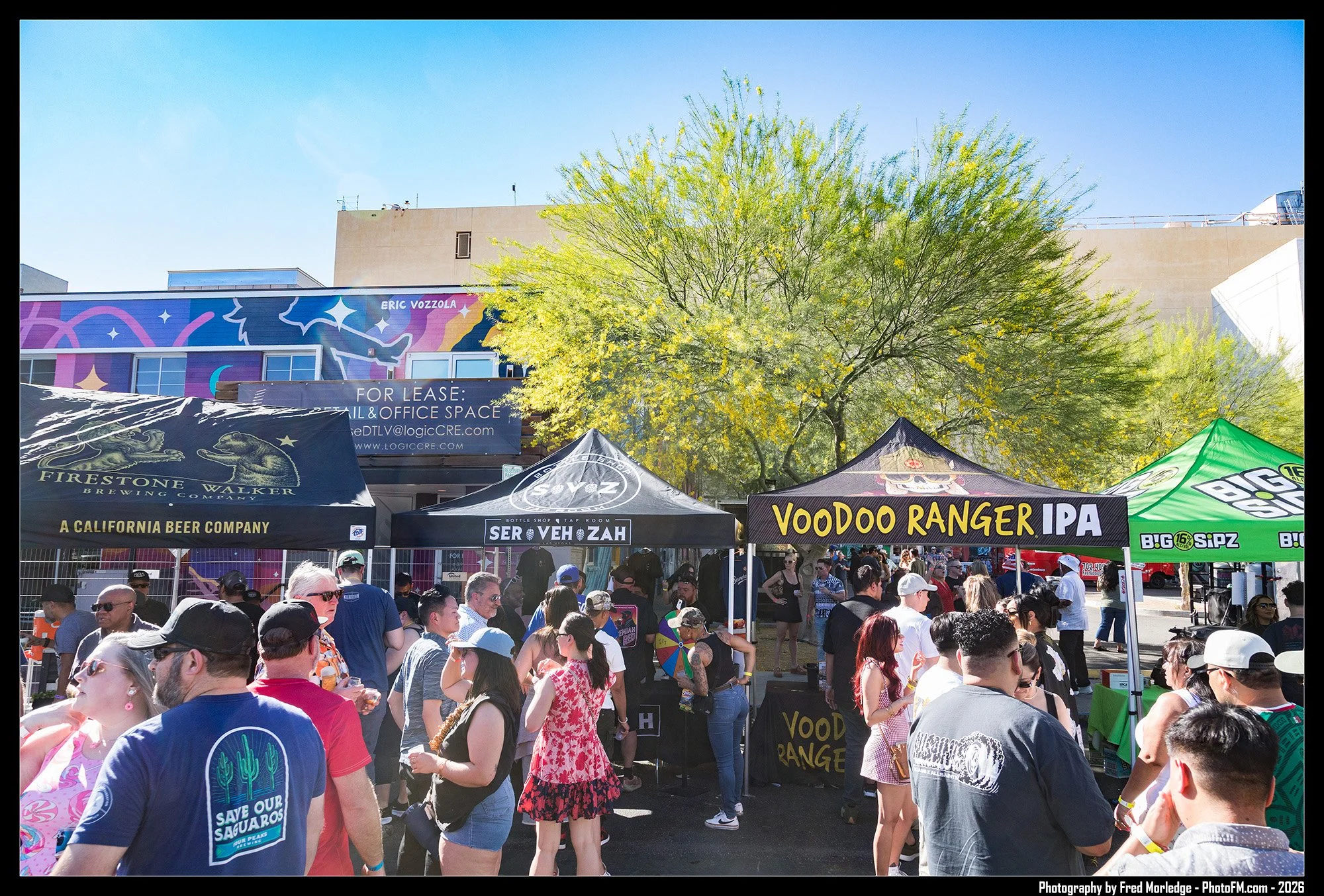 A crowded outdoor event with several vendor tents, including brands like Firestone Walker Brewing Co., Serv zah, and Voodoo Ranger IPA. People are mingling under a clear blue sky, with a large green tree providing shade and a mural on a building in t
