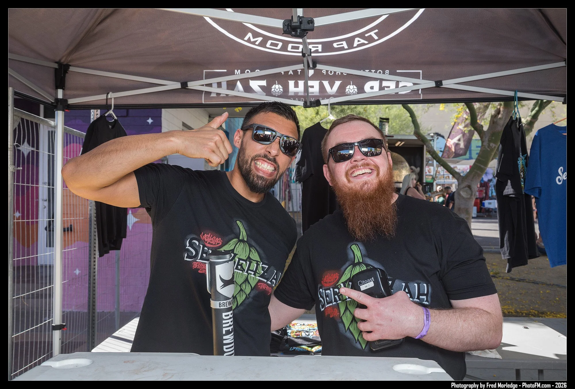 Two men wearing sunglasses and matching black t-shirts with a green chili pepper design, smiling and posing for a photo under a canopy at an outdoor event. One man on the left is holding a black insulated tumbler, and the other on the right is pointi