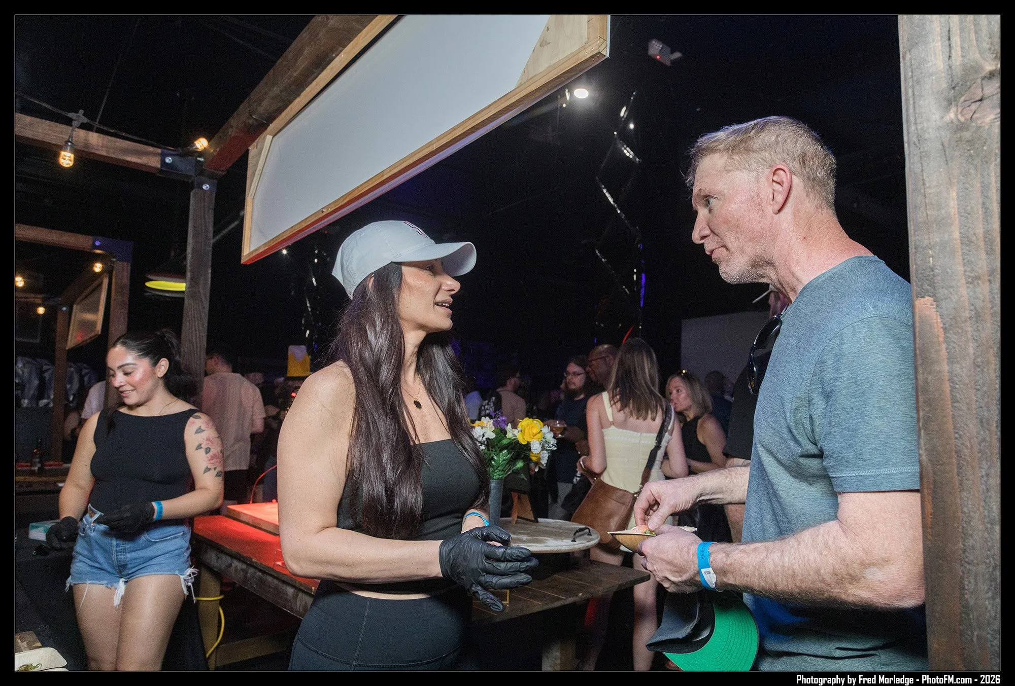 A woman with long dark hair wearing a white cap and black gloves hands a plate of food to a man in a blue t-shirt at a crowded indoor event. Other people are visible in the background, and a woman in a black top and denim shorts is seen to the left.