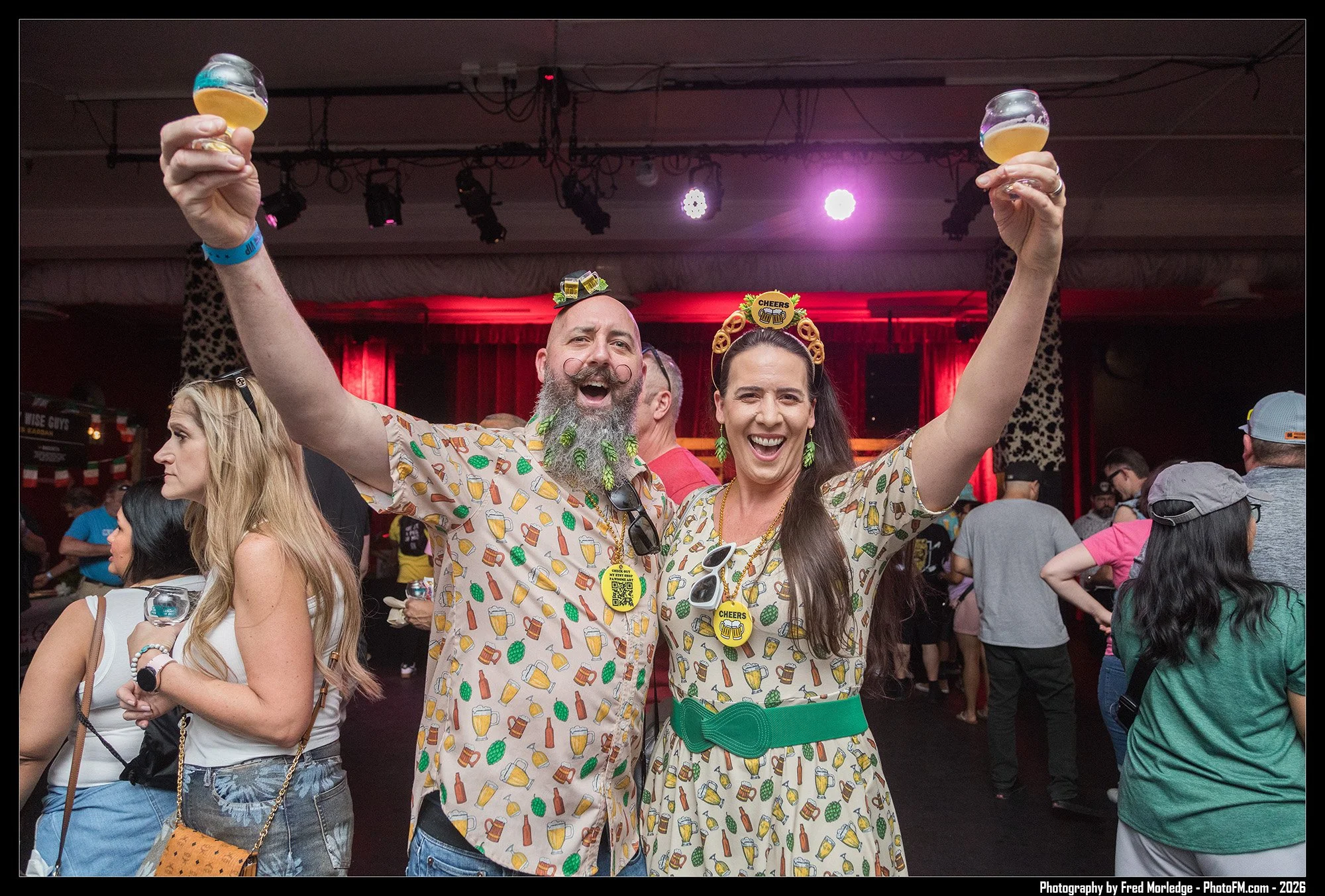 A group of people at a lively party or event. Two individuals in the foreground are wearing matching outfits with alcohol-themed patterns, celebrating with drinks raised in the air and wearing fun hats and accessories. The background shows other atte