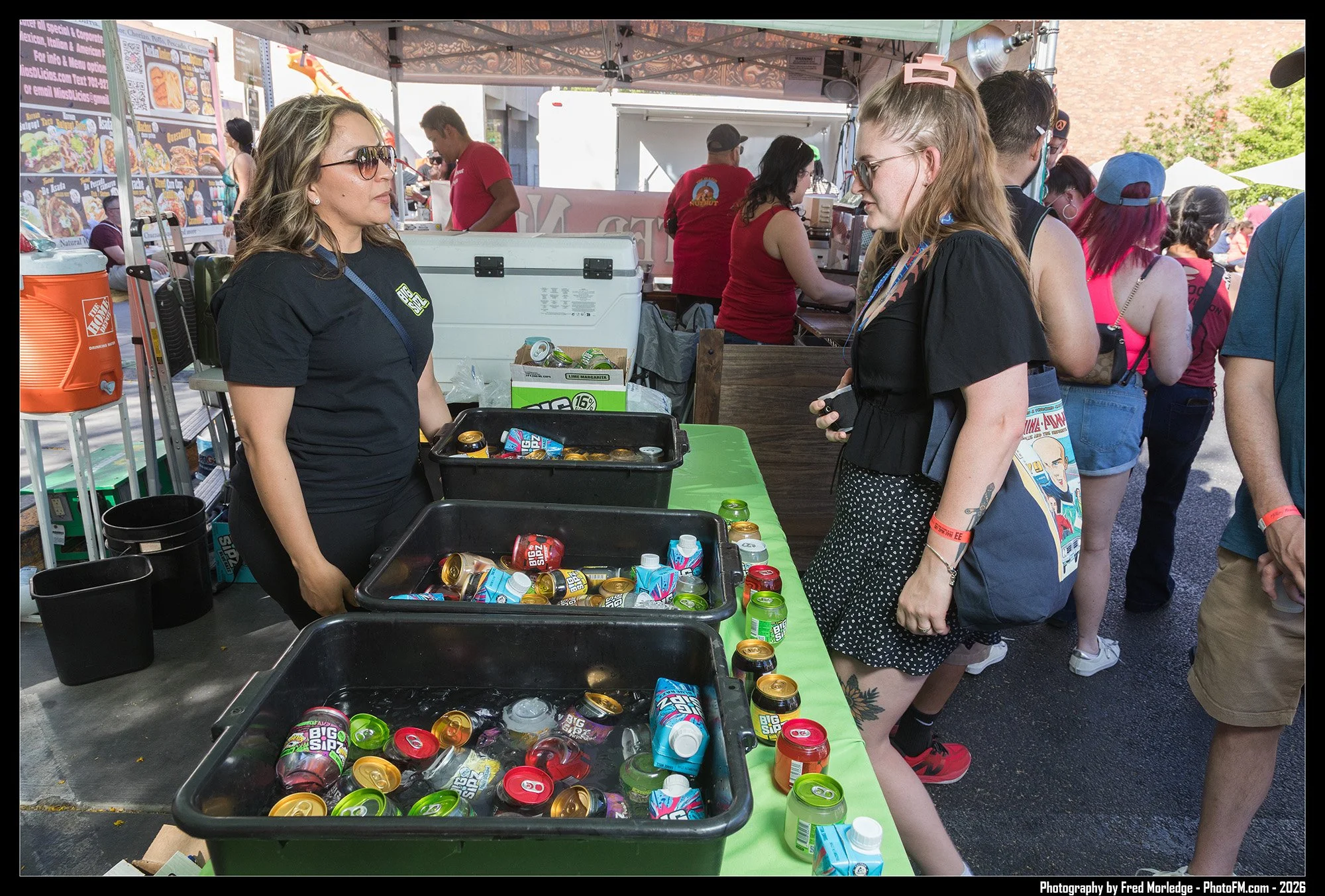 People gathered at an outdoor event, buying canned beverages from a vendor under a tent with food menus displayed behind them.