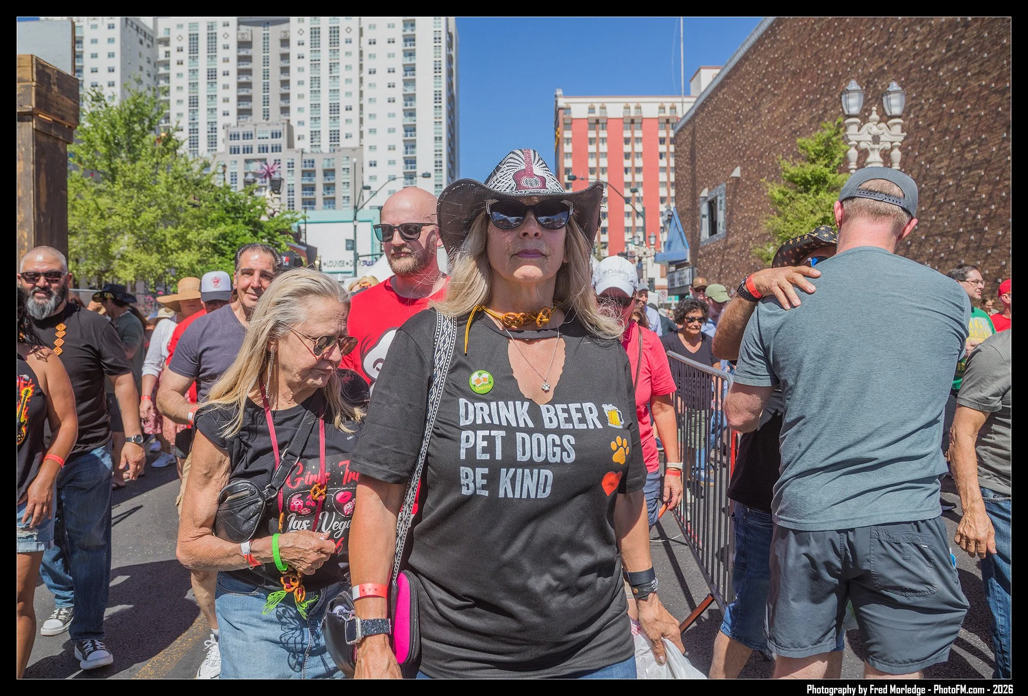 A group of diverse people attending a pride event on a city street, with tall buildings and blue sky in the background. The woman at the front wears sunglasses, a wide-brimmed hat, and a black T-shirt with a message about kindness and pet dogs. Other