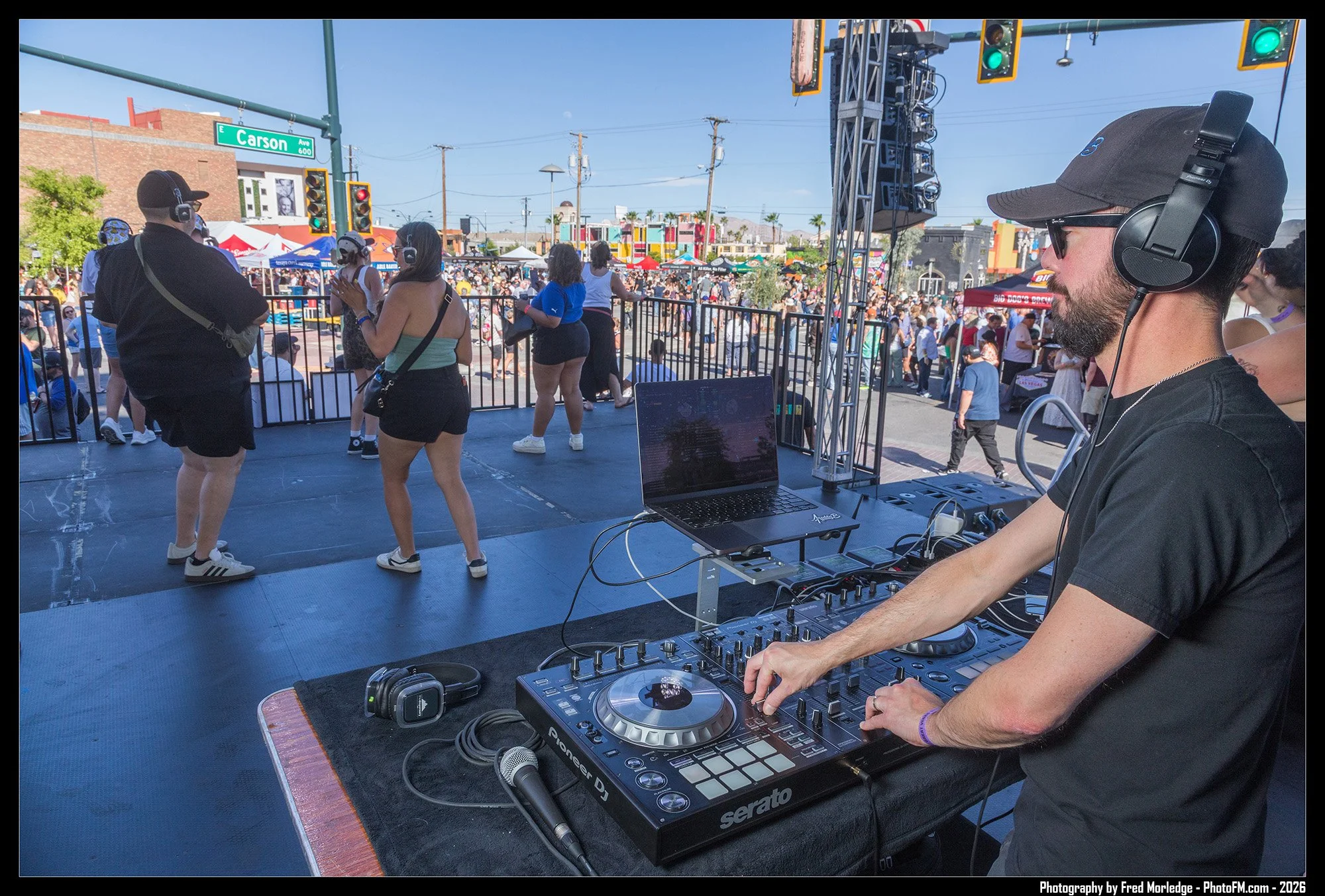 A DJ wearing headphones and sunglasses is mixing music at an outdoor event, with a crowd of people dancing and socializing in the background under a clear blue sky.