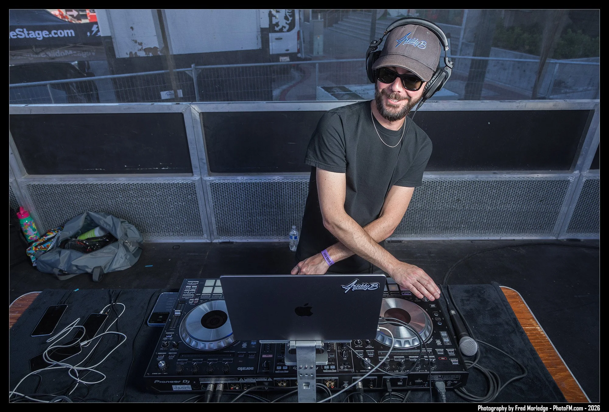 A DJ wearing sunglasses, a gray cap with 'Audio B' embroidered, and a black t-shirt, smiling at an outdoor event, standing behind a DJ setup with turntables, a laptop, and cables, with a metal barricade and a bus in the background.