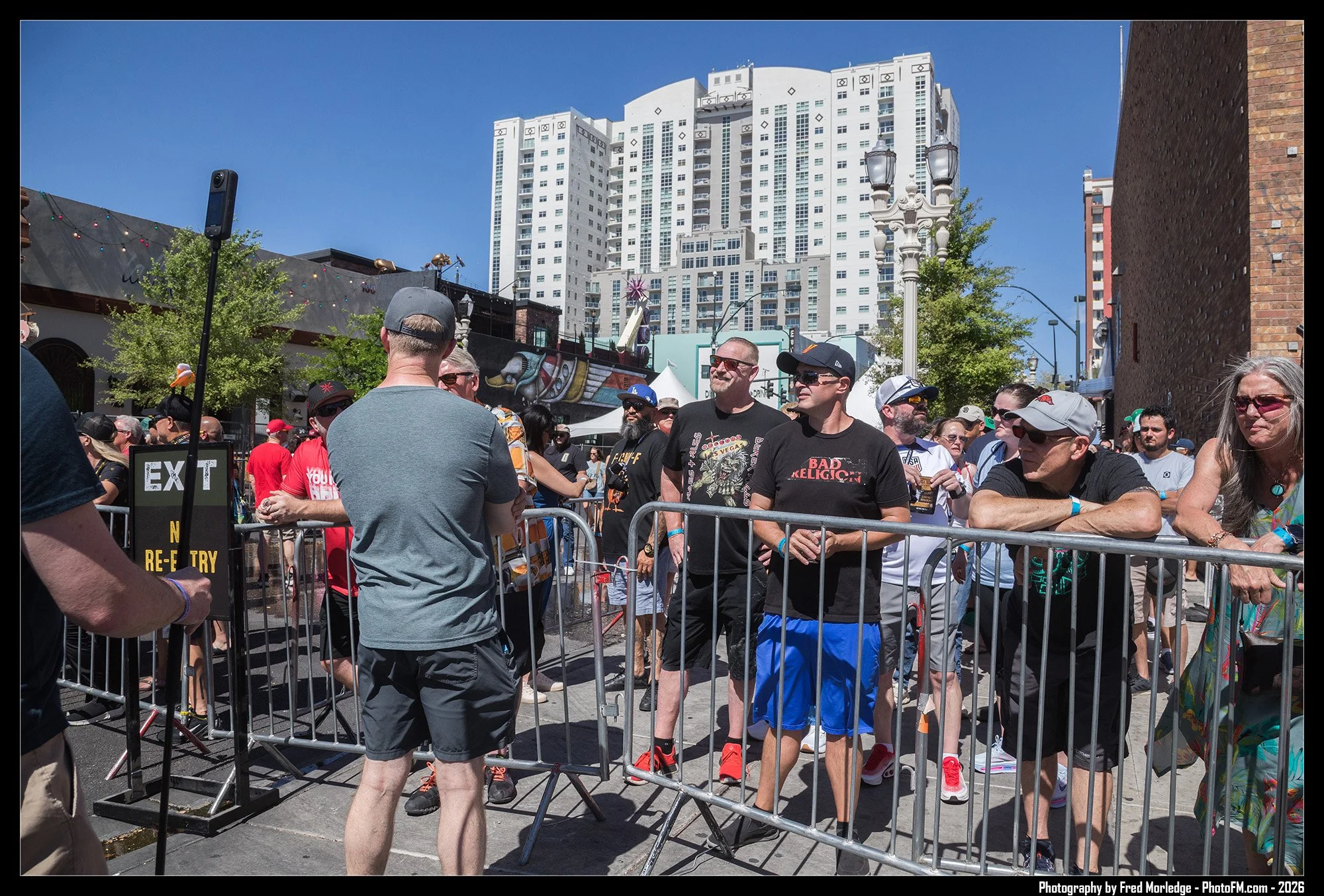 People waiting in line at an outdoor event, surrounded by metal barriers, with tall buildings and a clear blue sky in the background.