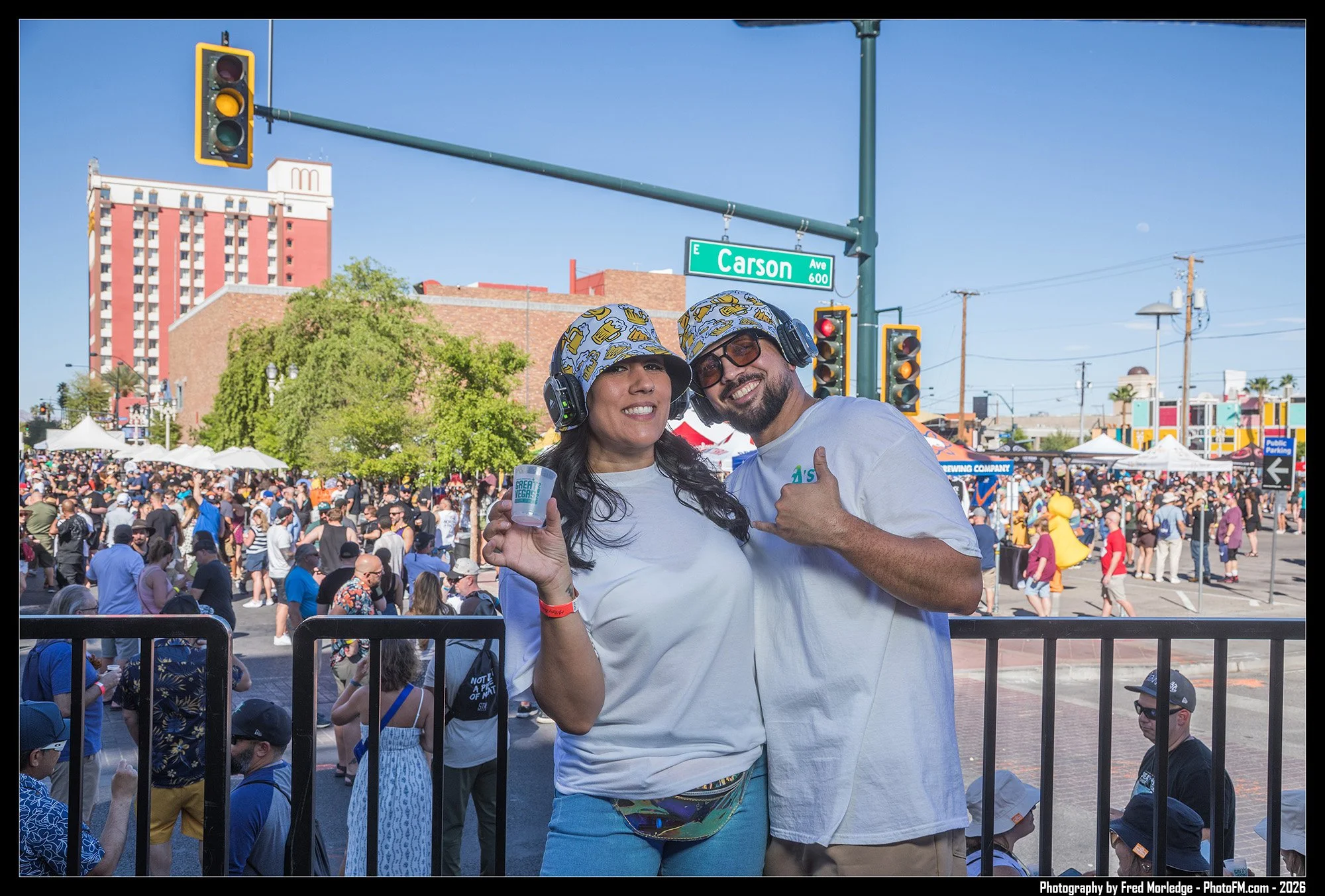 A smiling woman and man wearing matching hats and headphones at an outdoor event with a large crowd, street signs, and city buildings in the background.