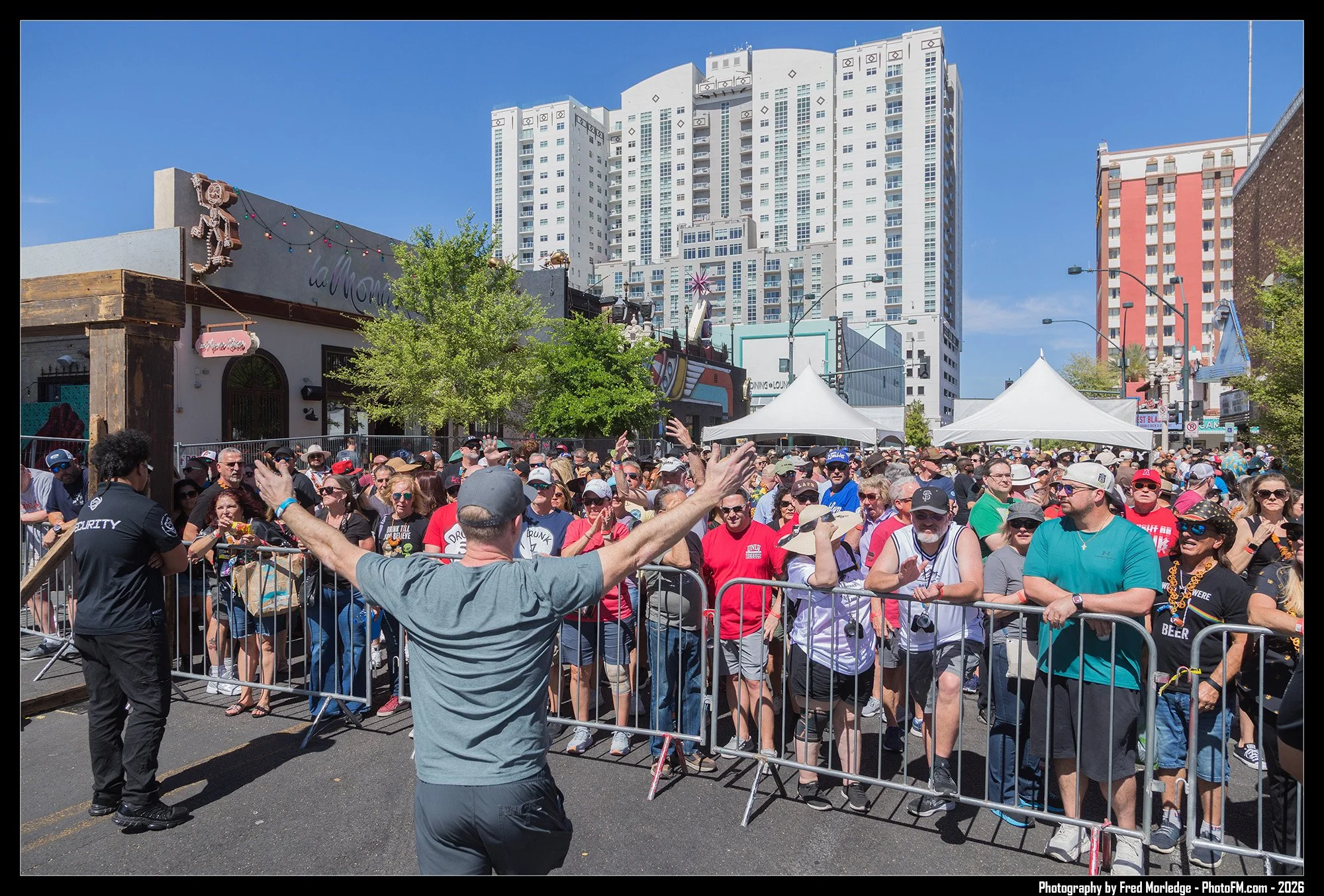 A crowd of people behind barricades during an outdoor event on a city street, with tall buildings and tents in the background.