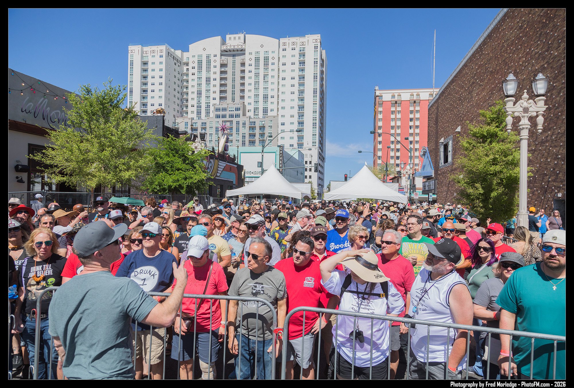 Crowd of people gathered in an urban outdoor area on a sunny day, with high-rise buildings in the background, some tents, and decorative street lamps.