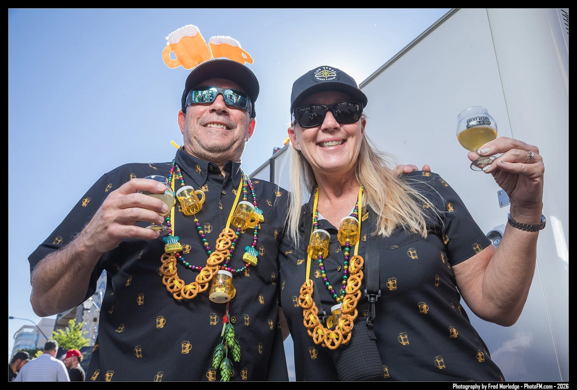 Two smiling adults, a man and a woman, celebrating at an outdoor event during the daytime. They are wearing black shirts with beer mug patterns, colorful bead necklaces, and black sunglasses. The man has beer mug headband, and they are holding glasse