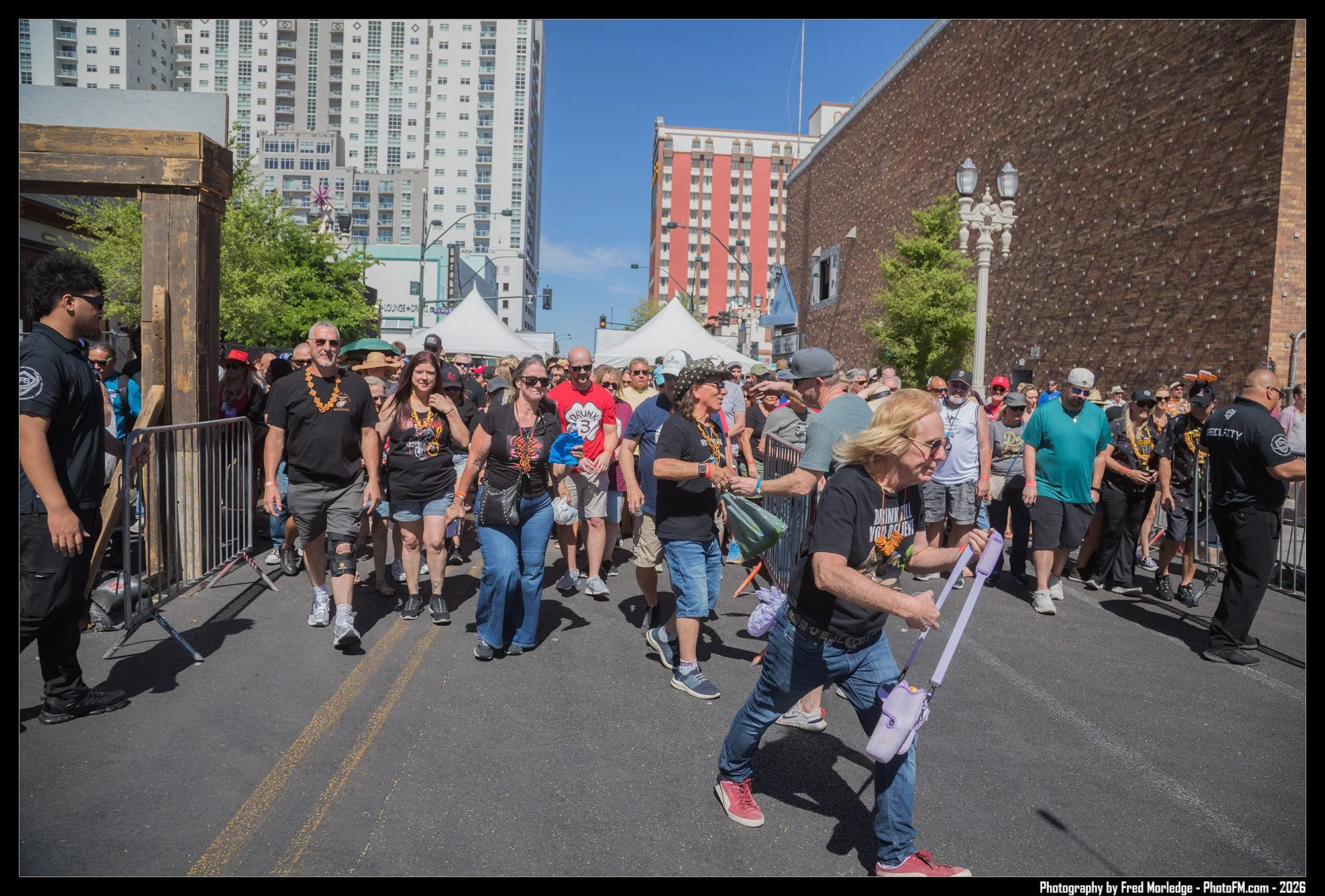 A crowd of people walking in a city street during a street festival or parade on a sunny day, with tall buildings in the background.