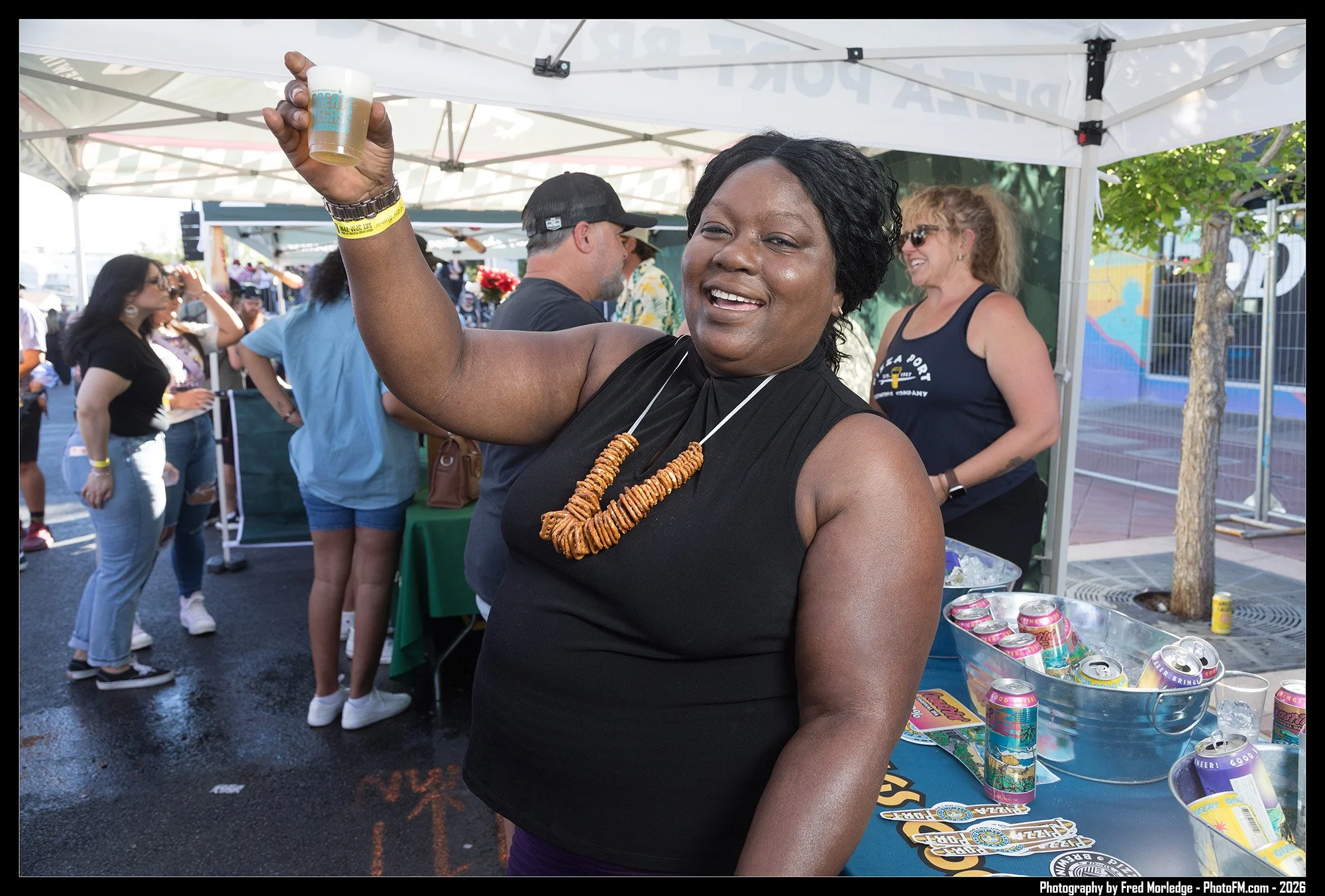 A woman wearing a black sleeveless top and a necklace of snack sticks, smiling and raising a beer, at an outdoor event under a white canopy with other people in the background.