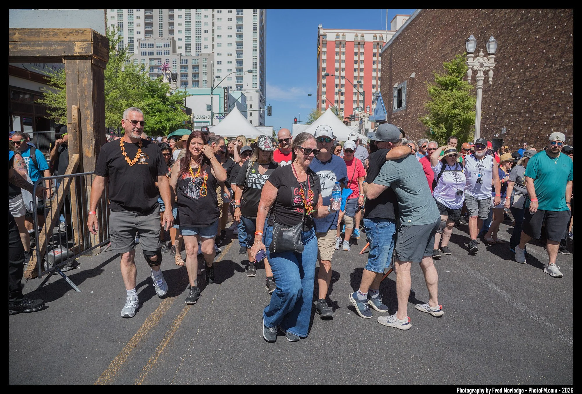 Crowd of people walking outdoors on a city street, with high-rise buildings in the background and tents set up nearby.