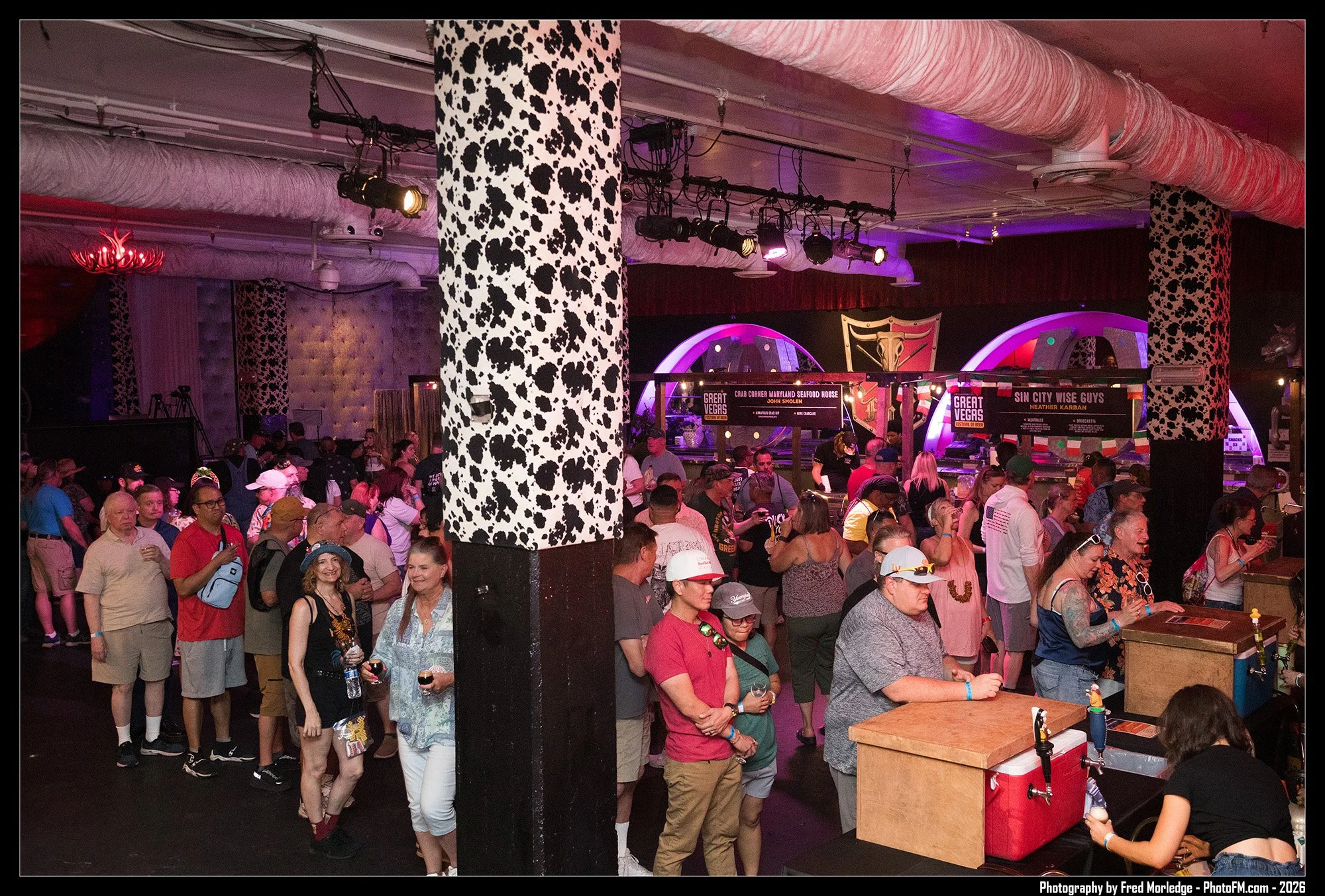 Crowd of people standing in line inside a venue with pink and purple lighting, decorated with cow-print patterned columns, overhead stage lighting, and signs for food stands.
