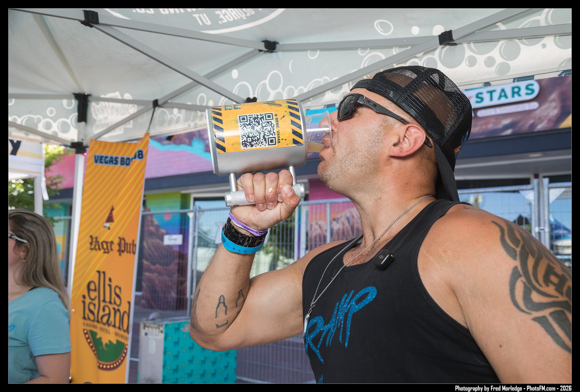A man wearing a black cap and sunglasses is drinking from a large cup with a QR code on it at a festival or fair. The background shows booths and colorful banners, including one for Vegas Island.