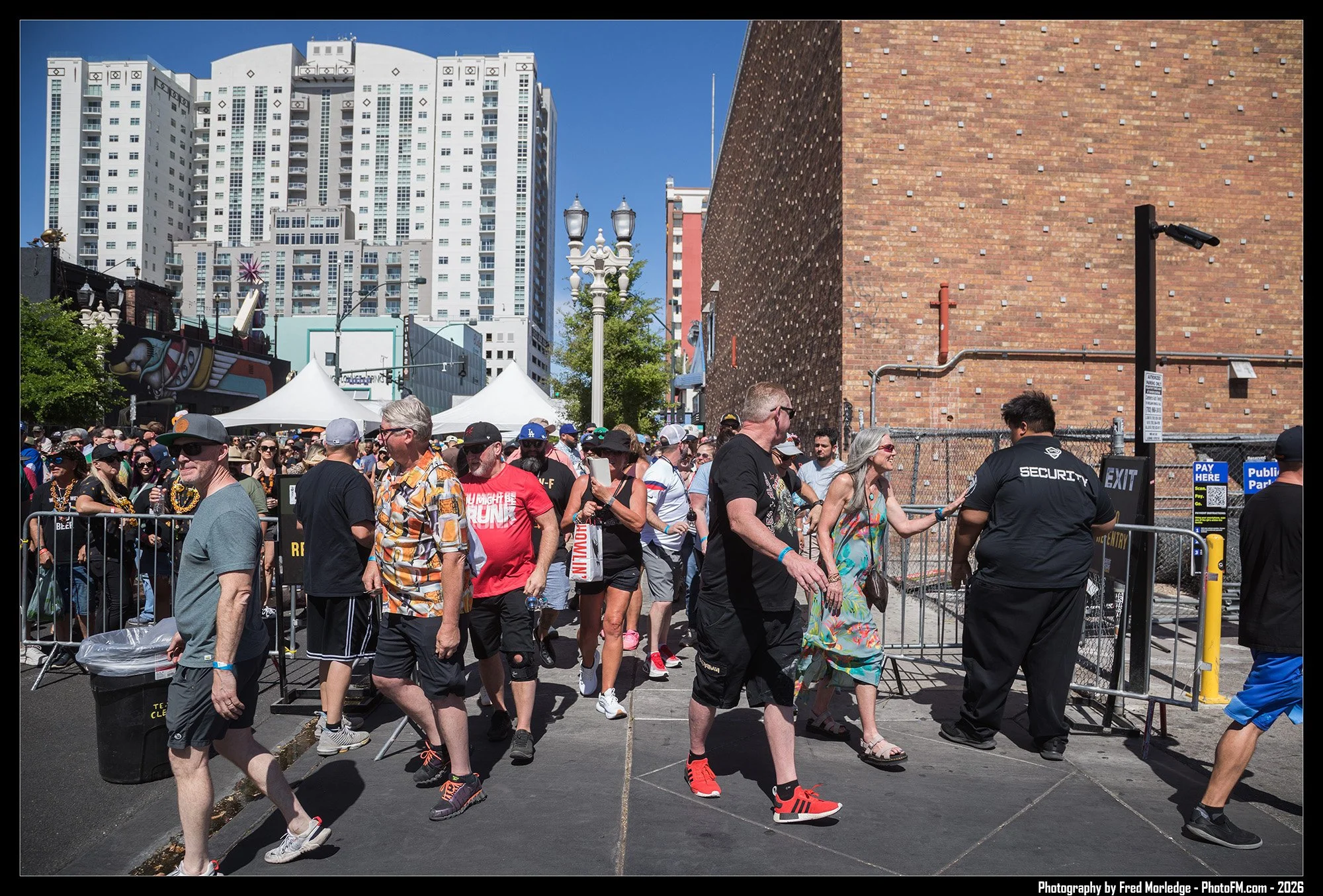 A crowd of people walking through a city street on a sunny day, with tall buildings in the background and security personnel at the entrance.