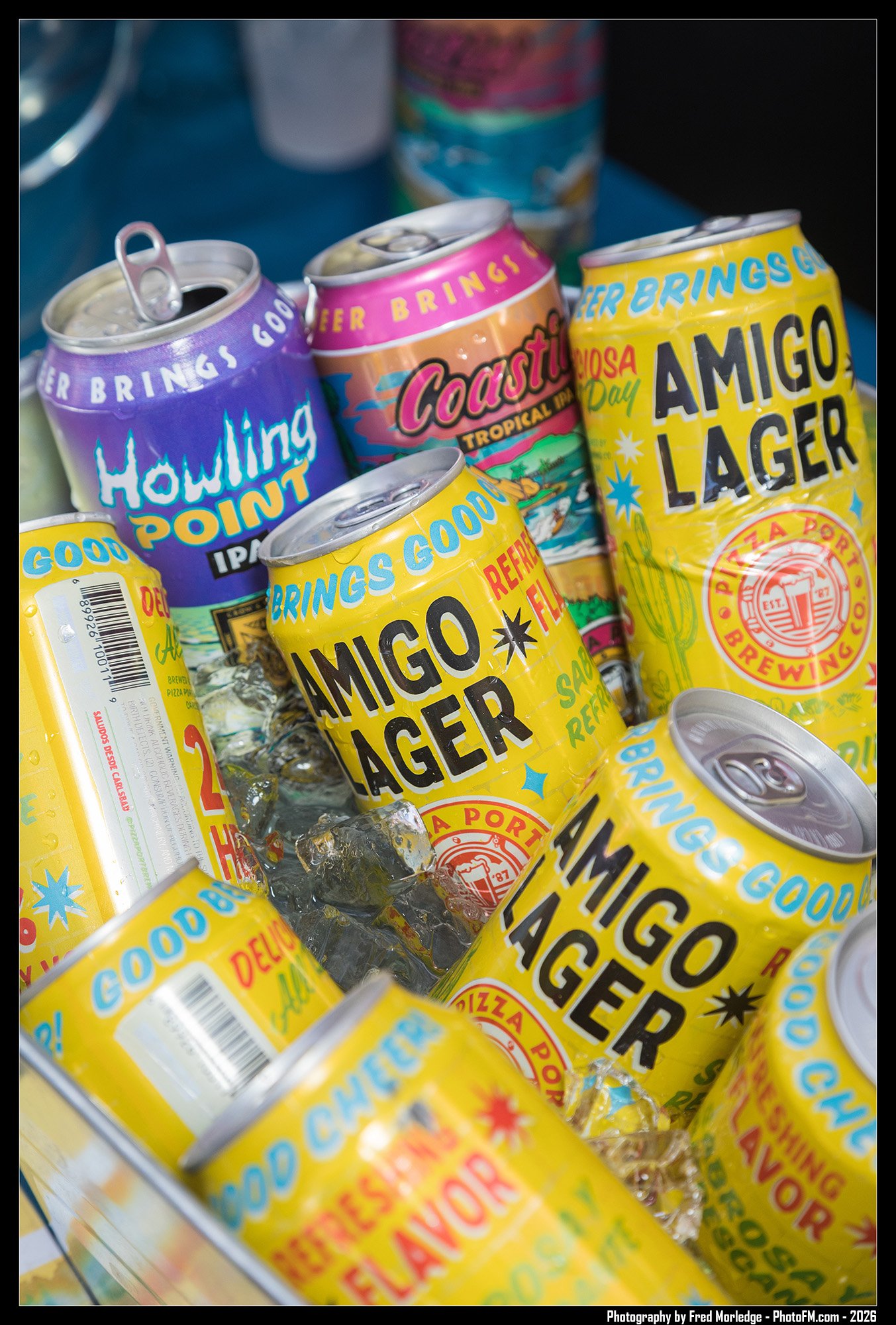 Several cans of Amigo Lager beer with colorful labels sitting on ice in a cooler. The cans feature bright yellow, purple, pink, and blue colors with various text and graphics.