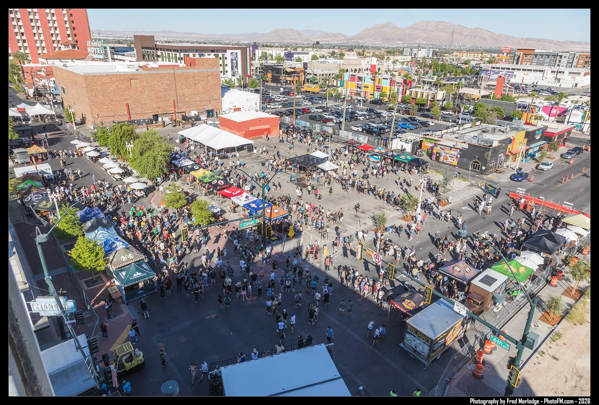 A crowded outdoor street fair in an urban area with vendor tents, food trucks, and many pedestrians walking along the street on a clear, sunny day. In the background, there are colorful buildings, parking lots, and mountains.