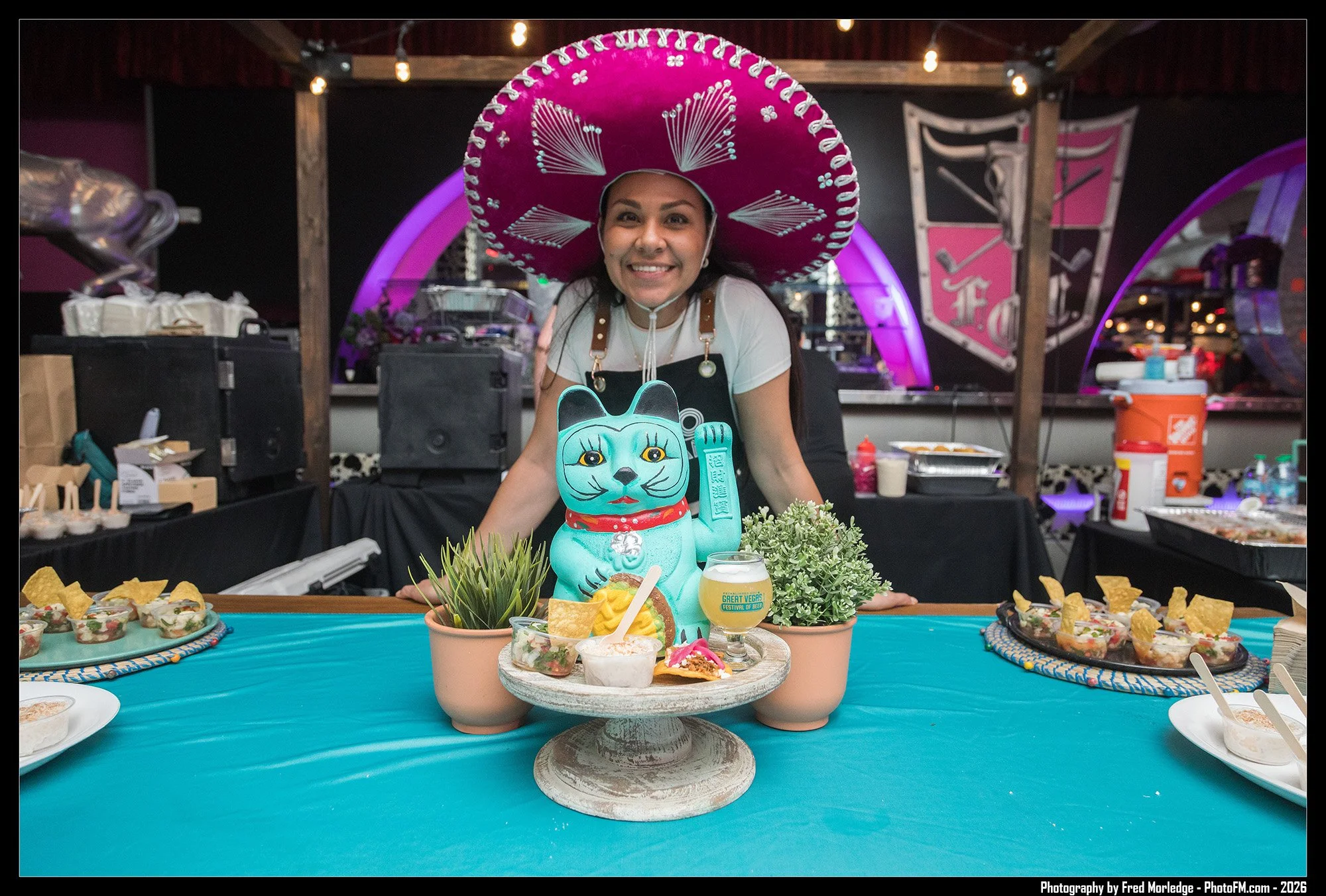 A woman wearing a large pink sombrero hat is smiling at an event table decorated with a teal tablecloth. The table displays a Maneki-neko cat figurine, small plants, and various plates of food. In the background, there are decorative arches and banne