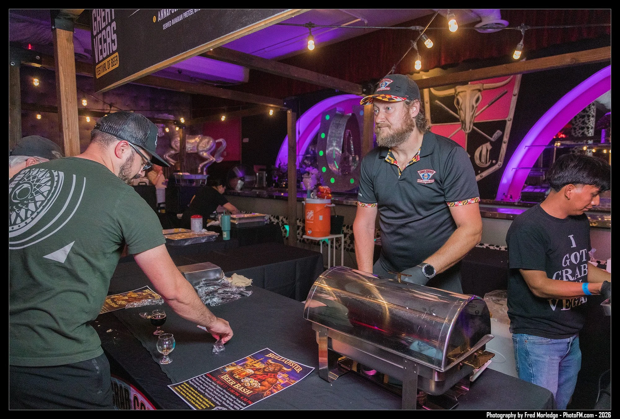 A group of people at an event, with one man wearing a baseball cap and black shirt, and another man with a beard, wearing a black shirt with colorful trim and a matching cap, standing behind a table with food and drink. There are posters and promotio