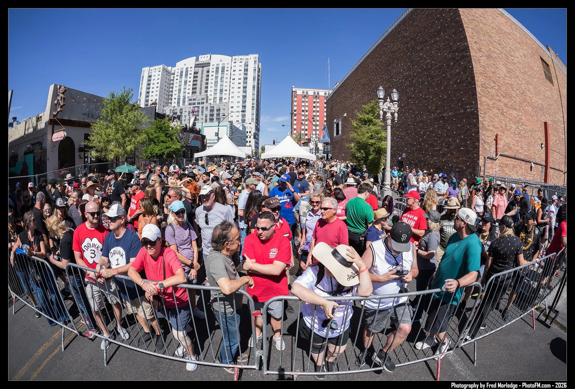 A large crowd of people standing behind metal barricades in an outdoor urban setting with high-rise buildings and tents in the background.