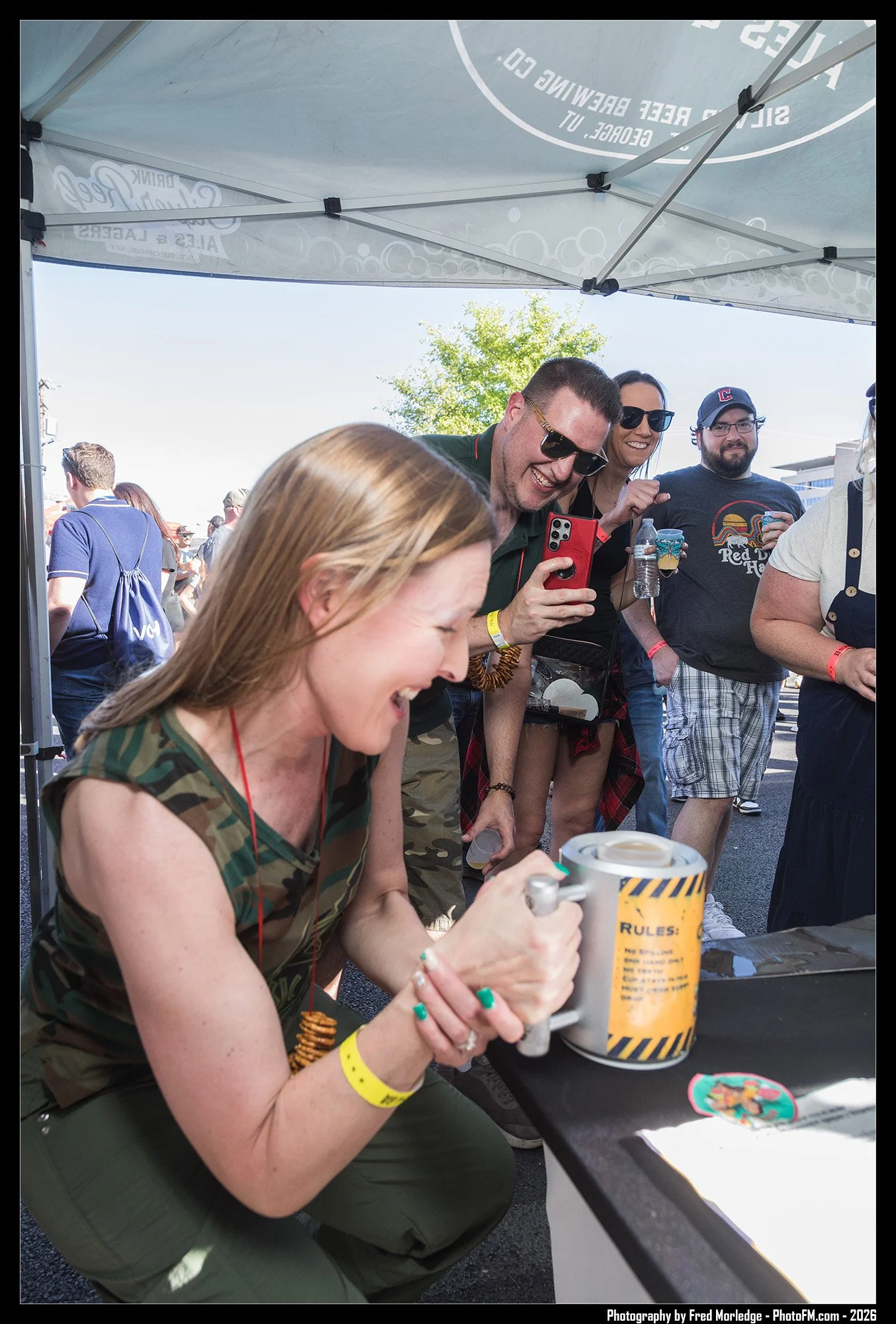 A group of people at an outdoor event, with two women at a table and several others in the background. The woman in the foreground is wearing a camouflage tank top and holding a cup, others are smiling and taking photos.