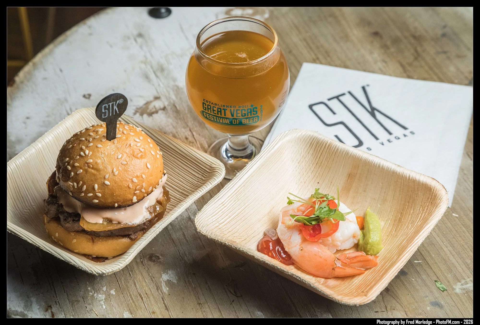 A burger with sesame seed bun, lettuce, and sauce, served in a disposable tray next to a seafood dish with shrimp, tomato, cilantro, and lime, with a glass of beer on a wooden table, with a napkin that reads "STK" and "Great Vegas Festival of Beer".