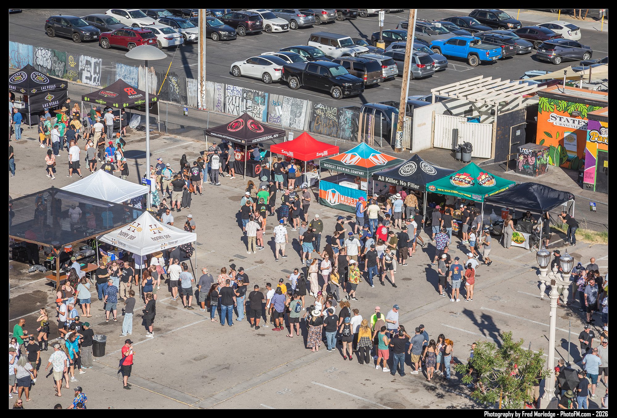 Large outdoor gathering with people lining up and walking in front of multiple vendor tents, in an urban parking lot area with parked cars and graffiti on the wall behind.