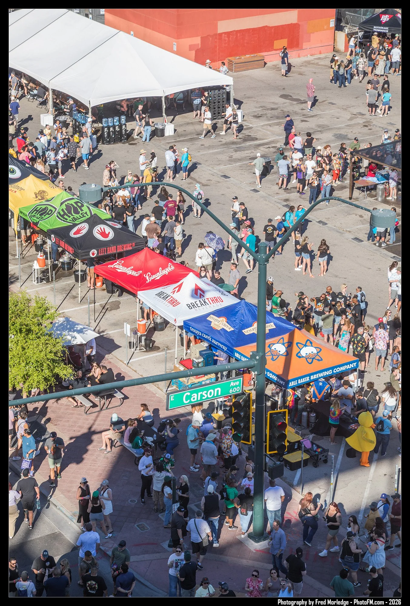 Crowd of people standing in line and socializing at an outdoor event on a city street, with vendor tents and a large white tent, and a street sign for E Carson Ave at the corner.