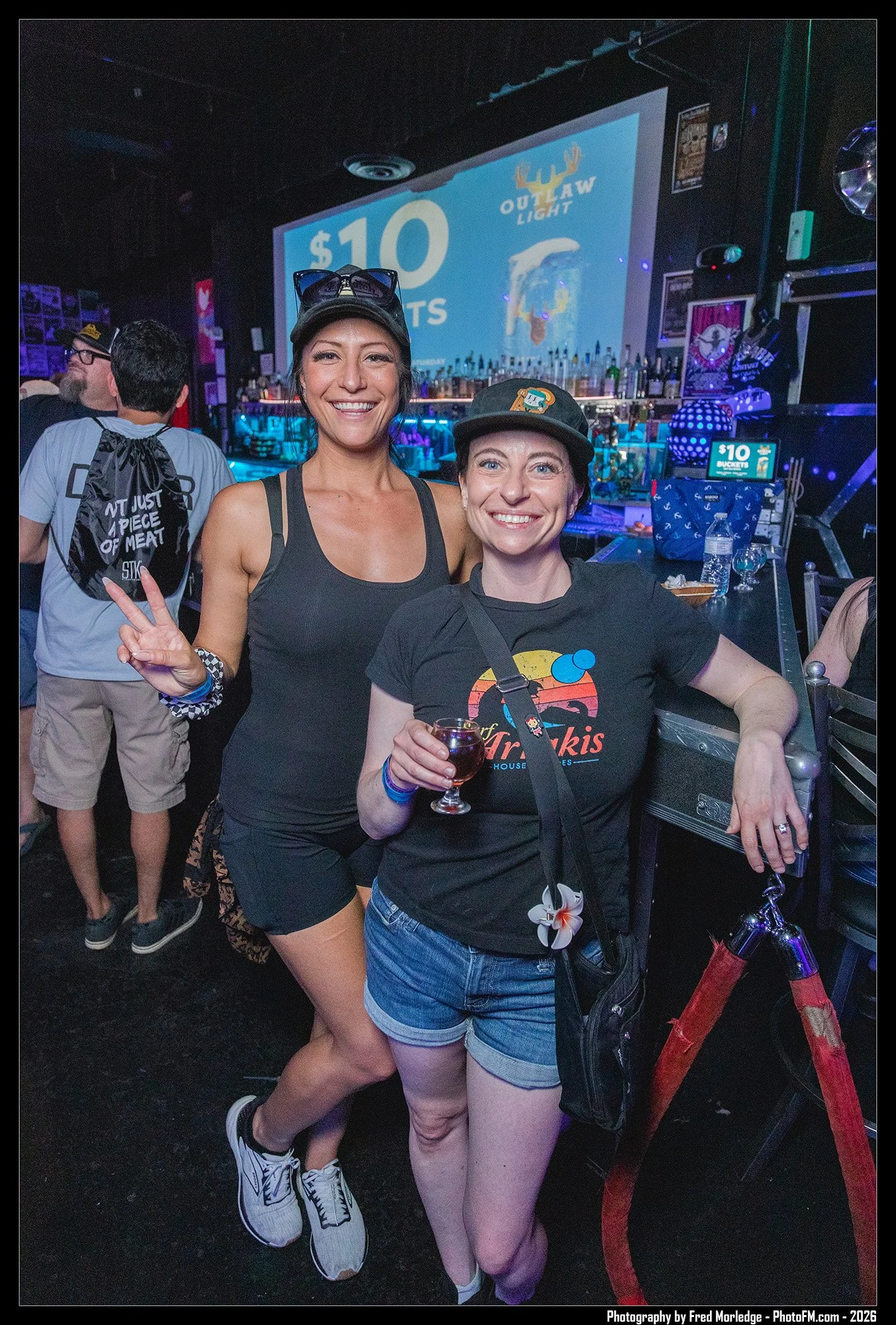 Two women smiling and posing for a photo inside a bar or nightclub. One holds a glass of wine, making a peace sign with her fingers. The background shows a bar with bottles and a large screen displaying a $10 event or promotion. Both women are wearin
