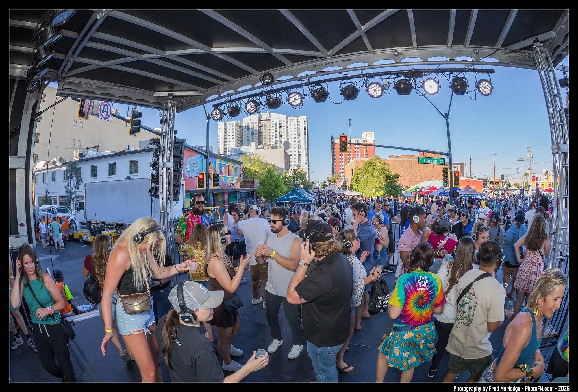 A large outdoor music event with many people gathered under a metal stage framework with stage lights. The crowd is diverse, wearing casual and colorful clothing, and some are wearing headphones. In the background, there are tall buildings and street signs, including one for Carson Avenue.