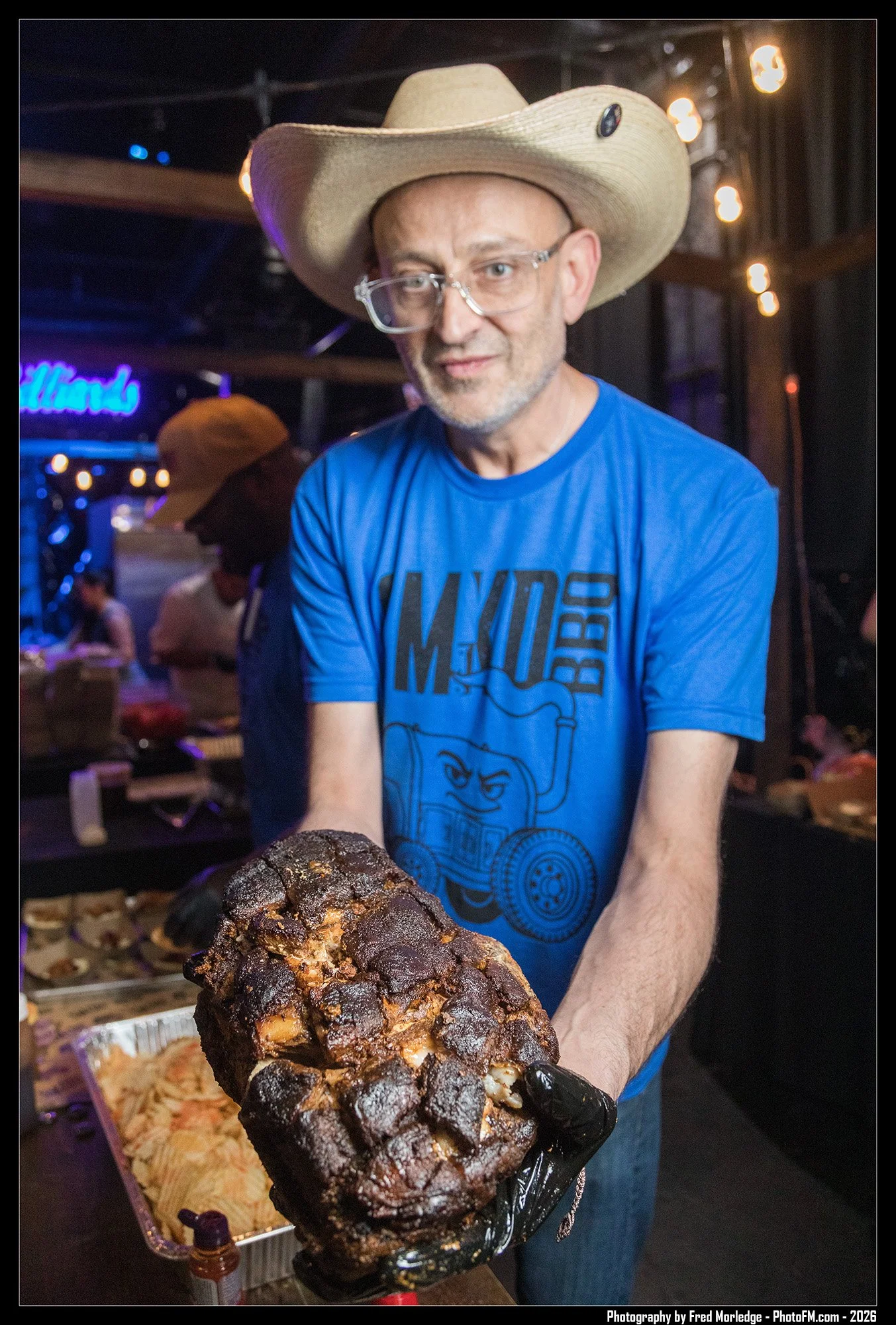 A man wearing a cactus-wrapped cowboy hat and glasses is holding a large, dark, charred-looking piece of meat at a food event or market. In the background, there are other people and food displays, with warm lighting and a neon sign.