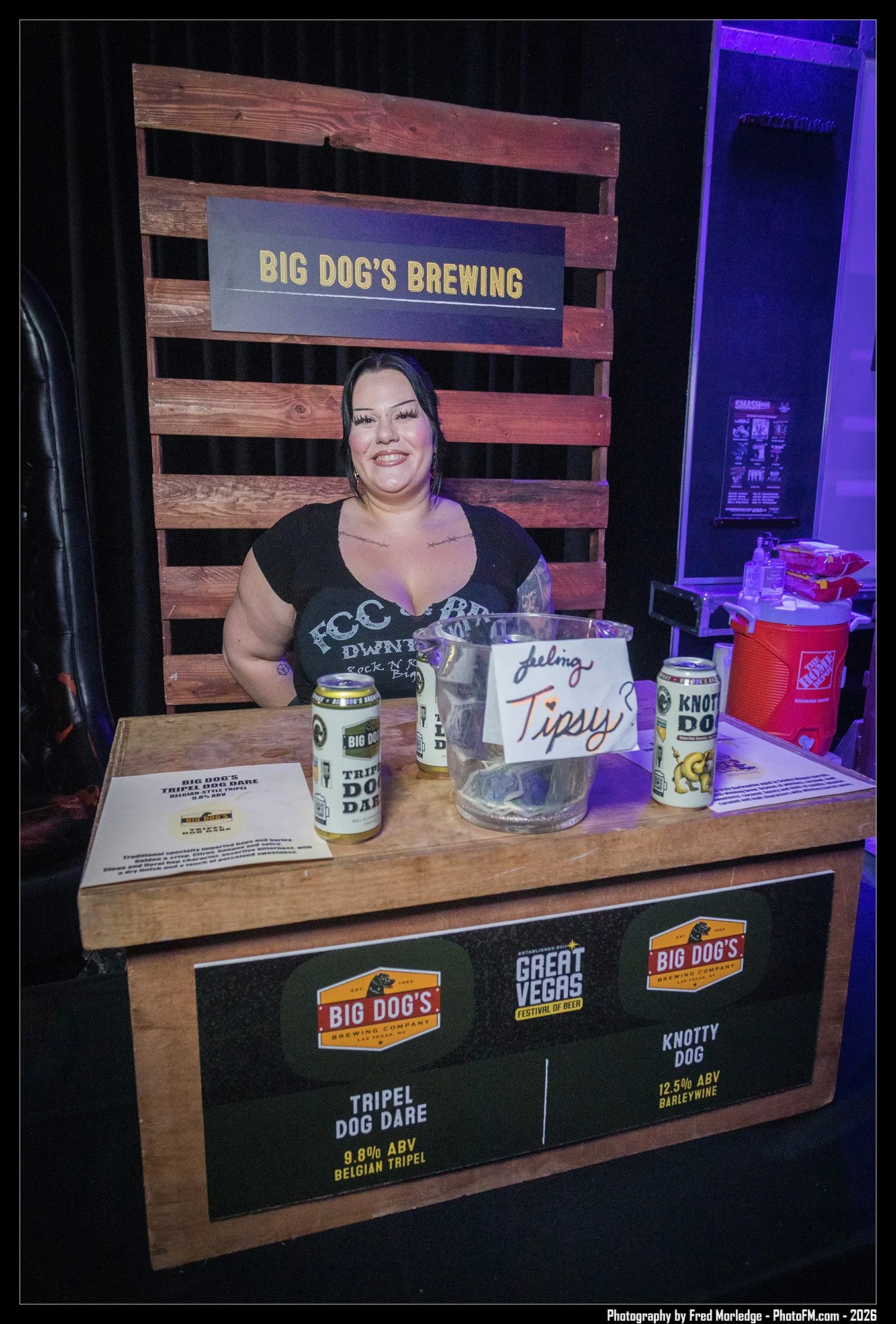 A smiling woman sitting behind a table at Big Dog's Brewing Company, with various beer cans and signs displaying beer names and alcohol content. A wooden backdrop with a sign reading "Big Dog's Brewing" is in the background.