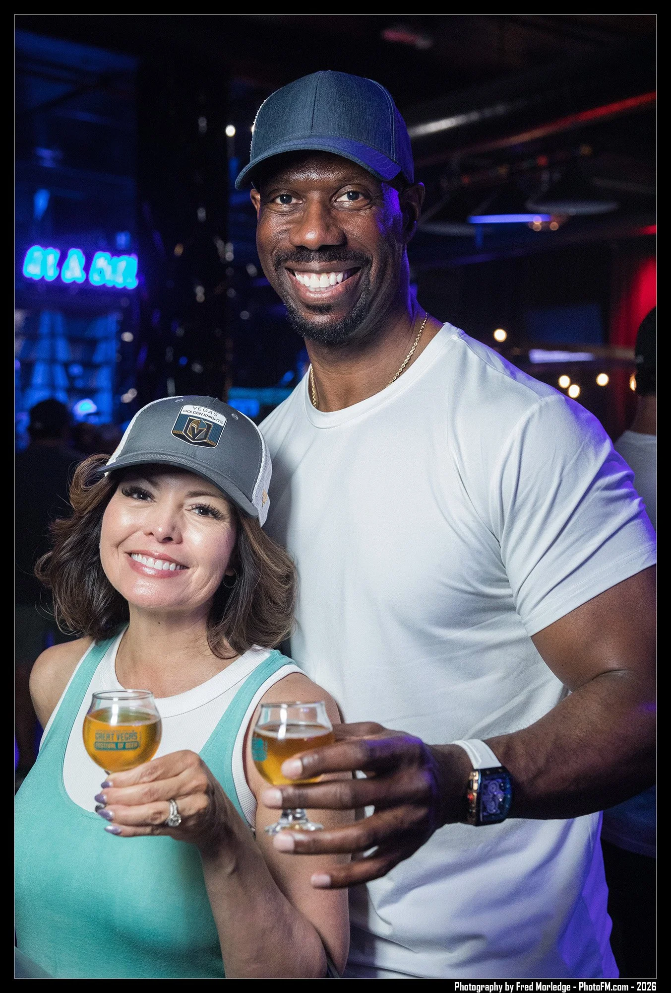 A group of three people at a bar or club, smiling and holding glasses of beer, with a neon sign in the background. Two women and one man, dressed casually, enjoying a social moment.