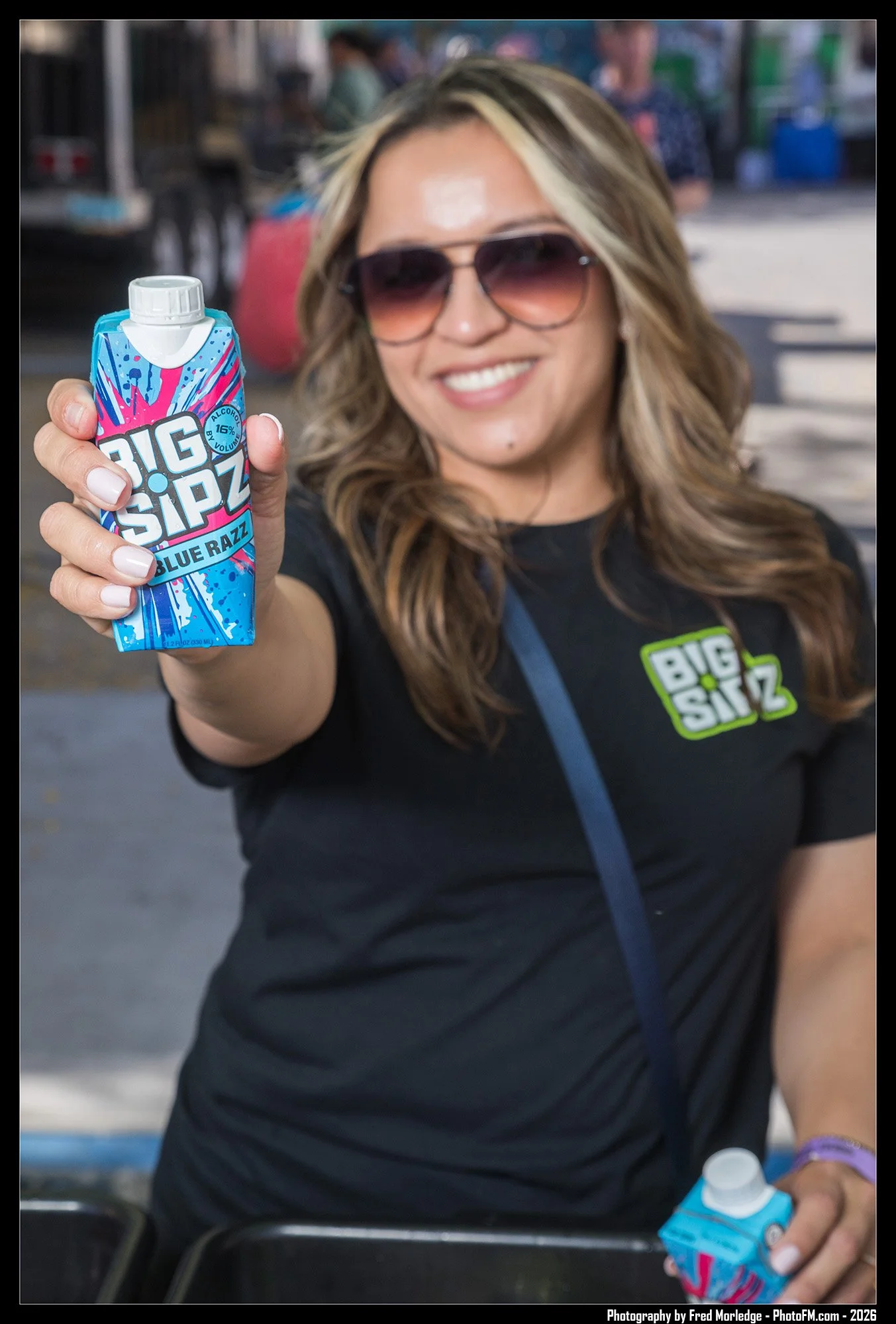 Woman wearing sunglasses and a black T-shirt holding a blue juice box with pink and blue design at an outdoor market or fair.
